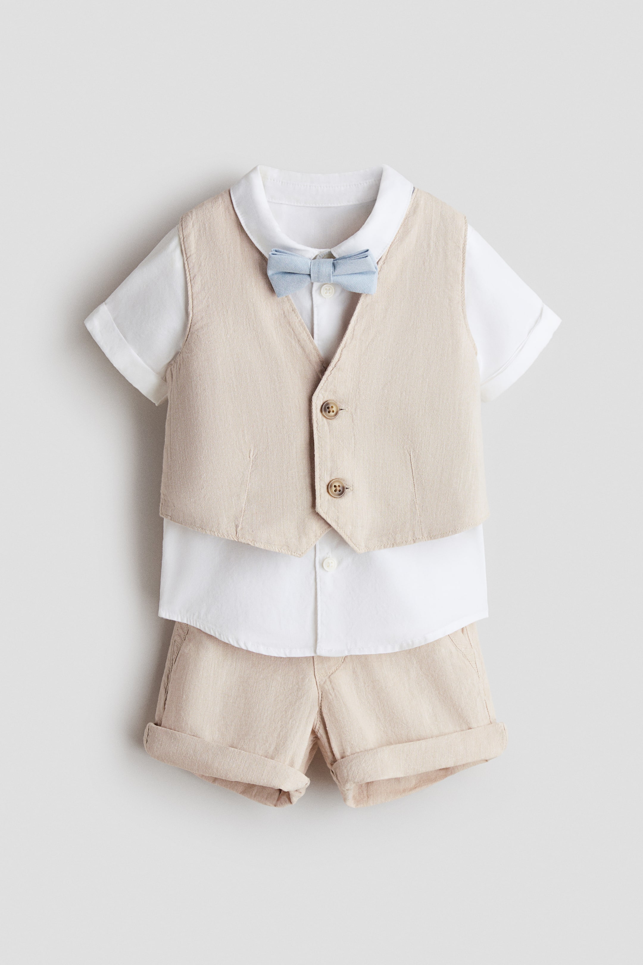 View larger image: A dressy set for a child is shown front-facing, featuring a light beige linen-blend vest with two visible brown buttons down the front, layered over a white short-sleeve cotton poplin shirt, and matching light beige linen-blend cuffed shorts with a decorative front pocket. A light blue pre-tied bow tie is worn at the shirt's collar.