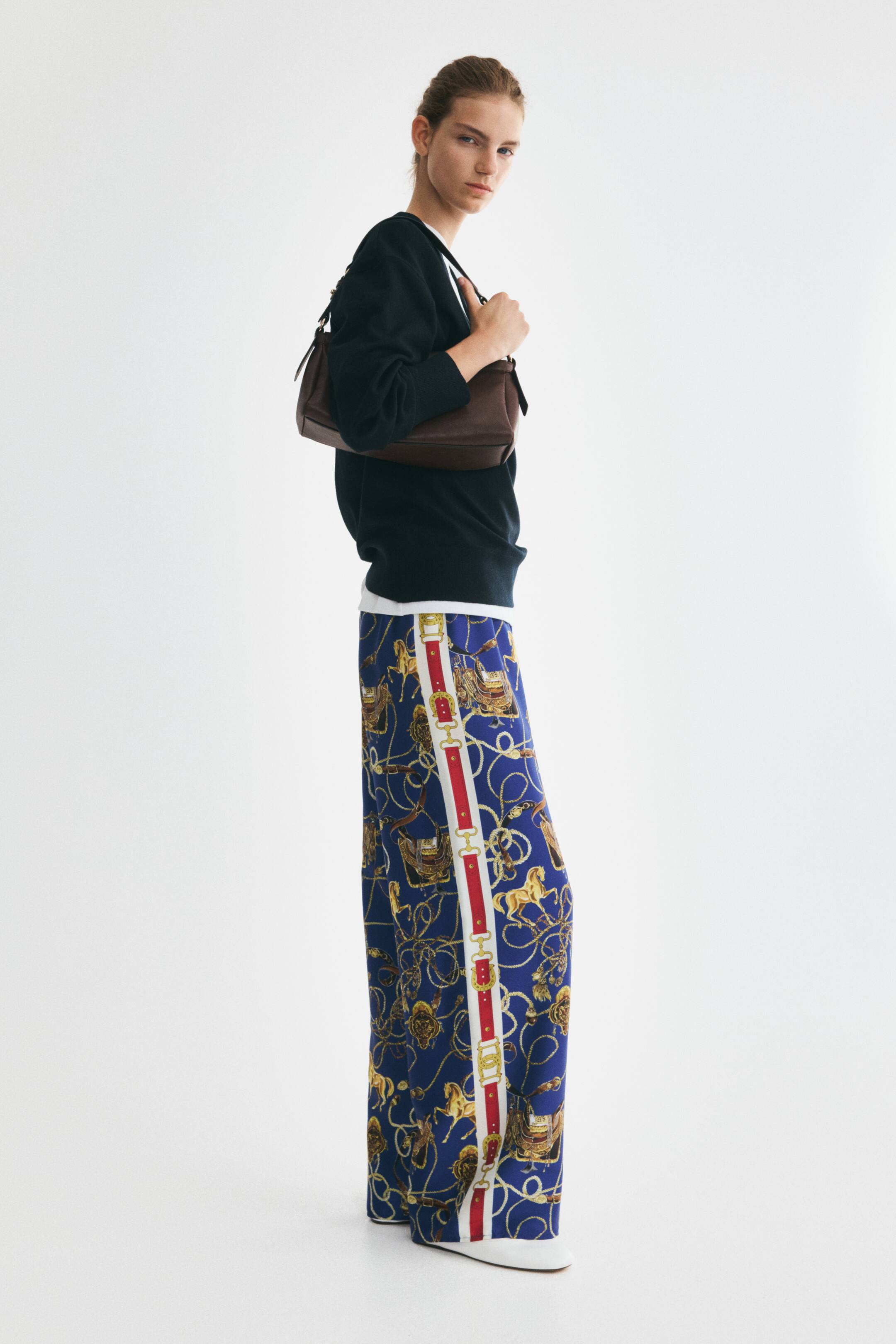 Vezi imaginea mai mare: Pantaloni cu croială relaxată, cu picior larg, din țesătură țesută cu aspect lucios, purtați de model. Pantalonii prezintă un imprimeu integral cu tematică ecvestră, în albastru și auriu, cu o dungă verticală roșie și albă și detalii aurii cu catarame de-a lungul părții exterioare a fiecărui crac.