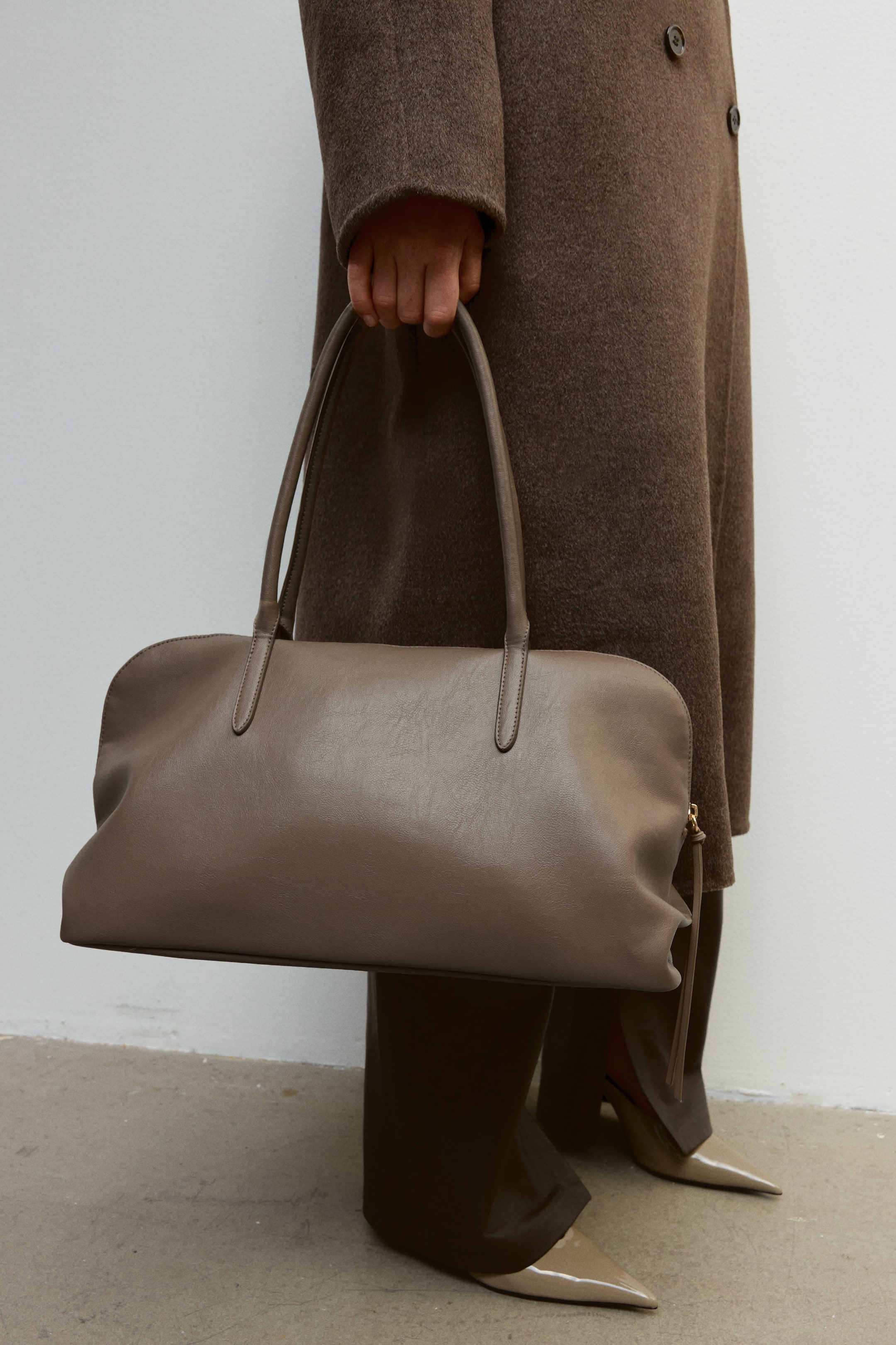 View larger image: A taupe leather handbag with dual slim top handles is being held by the model, who is styled in a long brown coat, dark trousers, and beige pointed-toe heels. The bag features a soft, rounded rectangular silhouette with a smooth finish.