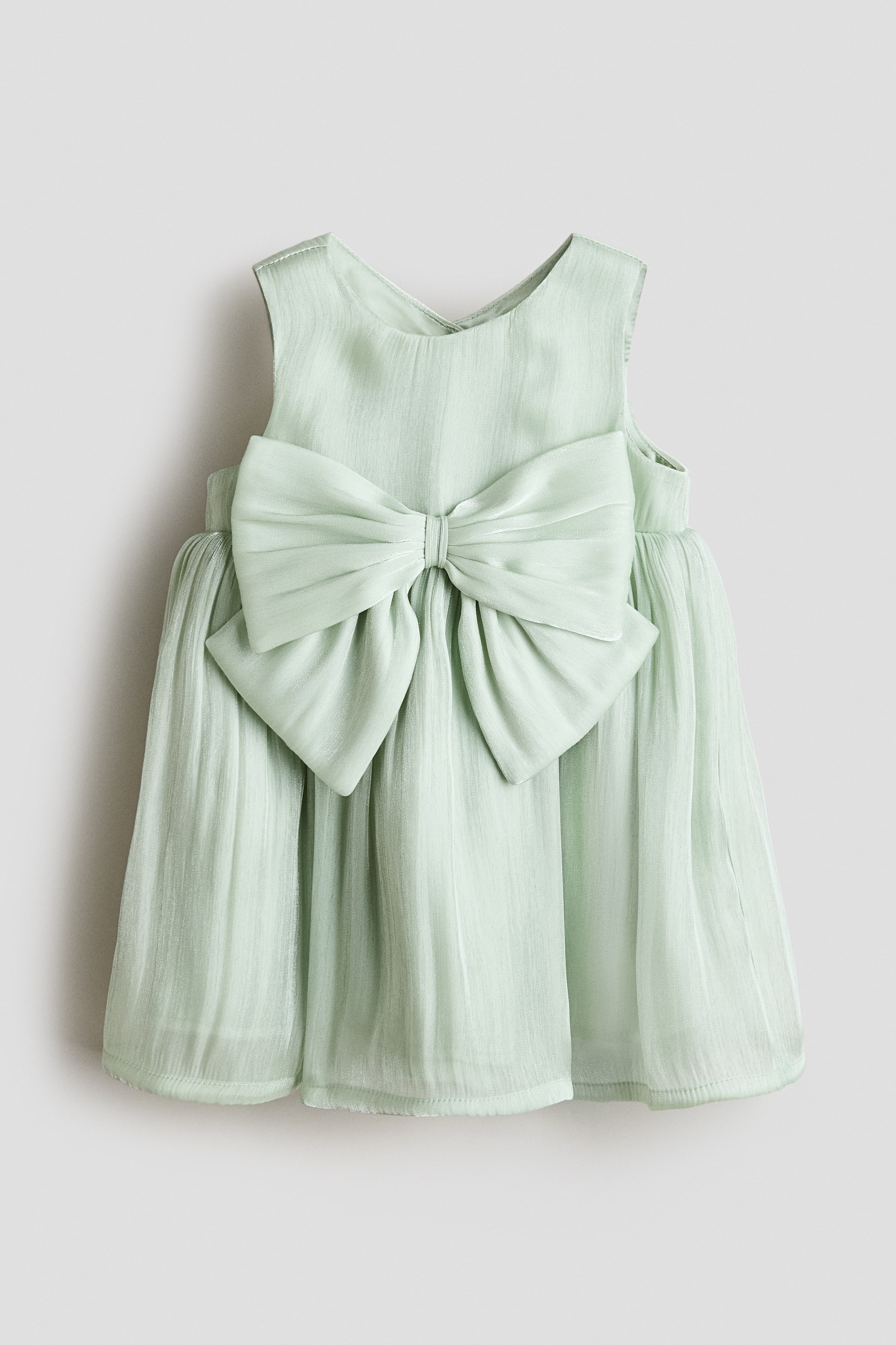 View larger image: A sleeveless dress in sheer organza is shown from the front, featuring a round neckline. A large, satin-trimmed bow adorns the front above the waist, where the dress gathers, creating a voluminous skirt.