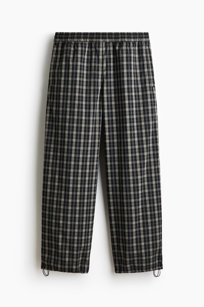 Trousers are shown from the front, showcasing a black and light brown check pattern. They feature an elasticated waistband and adjustable hems with visible drawstrings and cord locks at the ankles. - 1