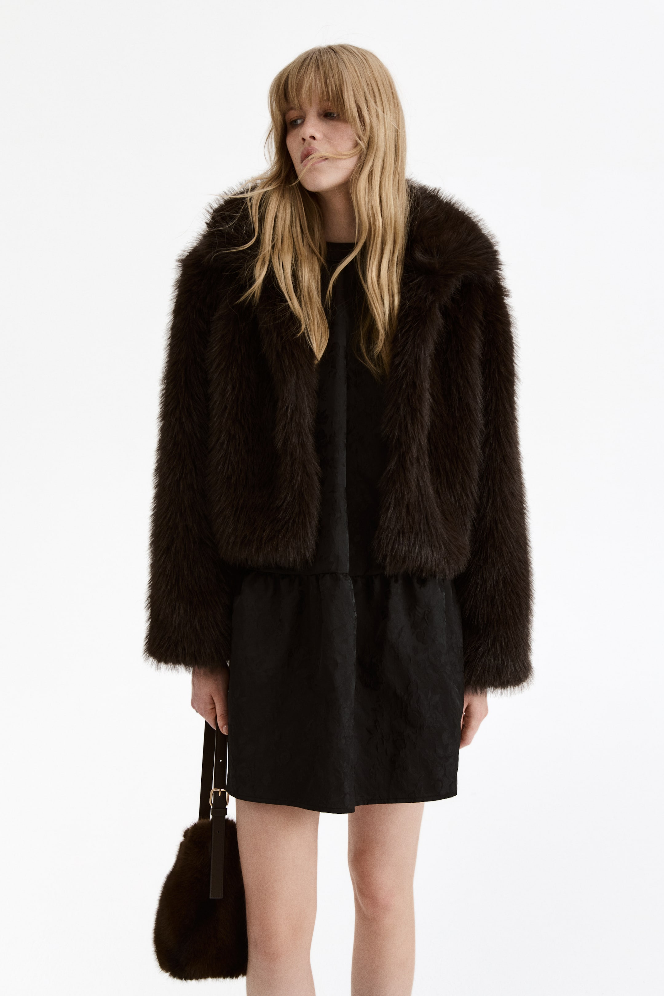 View larger image: A dark brown, cropped faux fur jacket is being presented front-facing, worn open over a black mini-dress that is featuring a subtle patterned texture and a gathered waistline. A matching dark brown faux fur bag with a slender strap is being held.