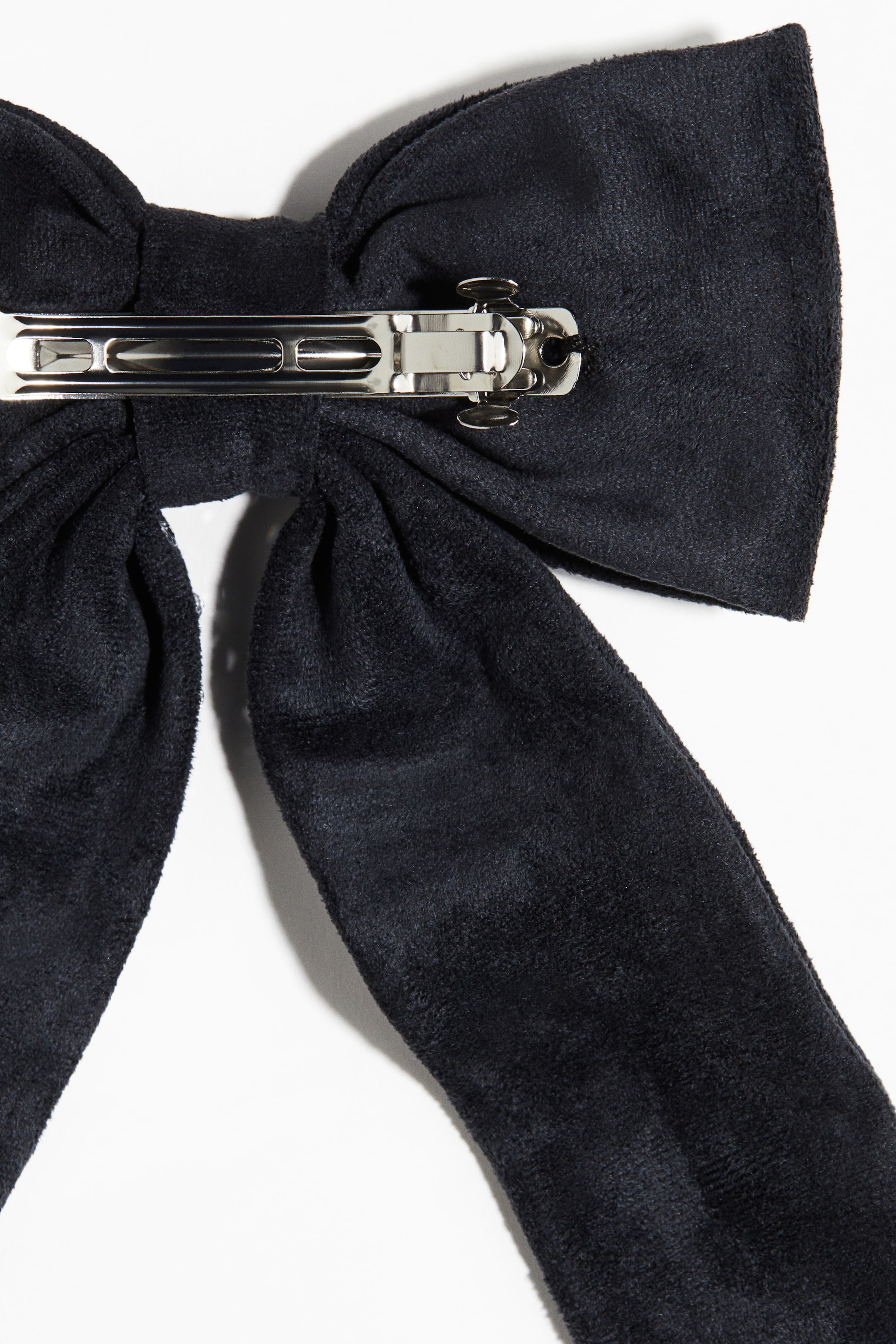 View larger image: A black hair bow is presented, featuring a plush, soft fabric forming its loops and flowing tails. A silver-toned metal barrette clip is visible on the back, positioned horizontally across the bow's cinched center and showing its spring mechanism.