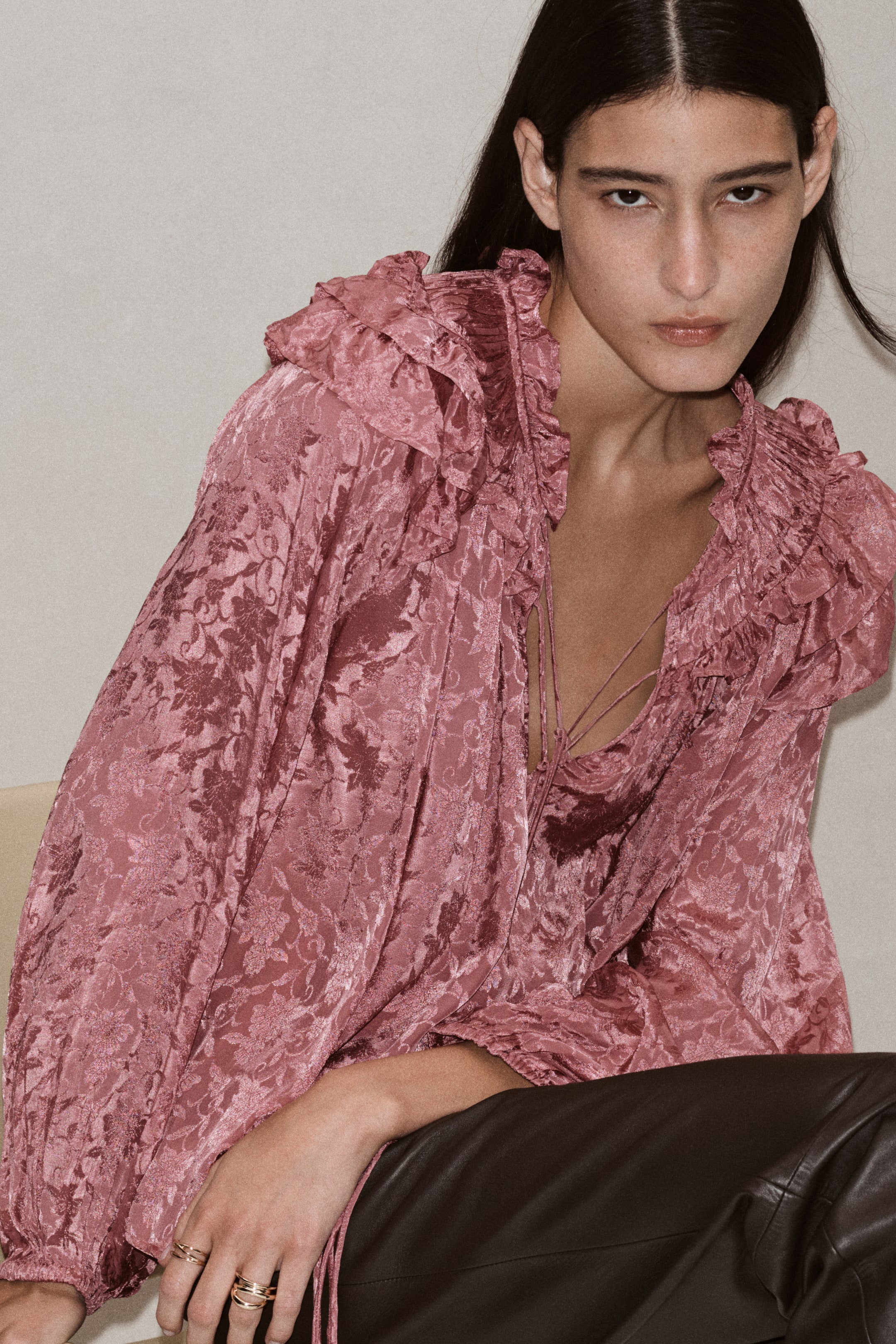Ver imagen más grande: La modelo lleva una blusa jacquard floral rosa con cuello con volantes, apertura frontal en V con lazos finos y mangas largas. La blusa se combina con un pantalón oscuro de corte estructurado.