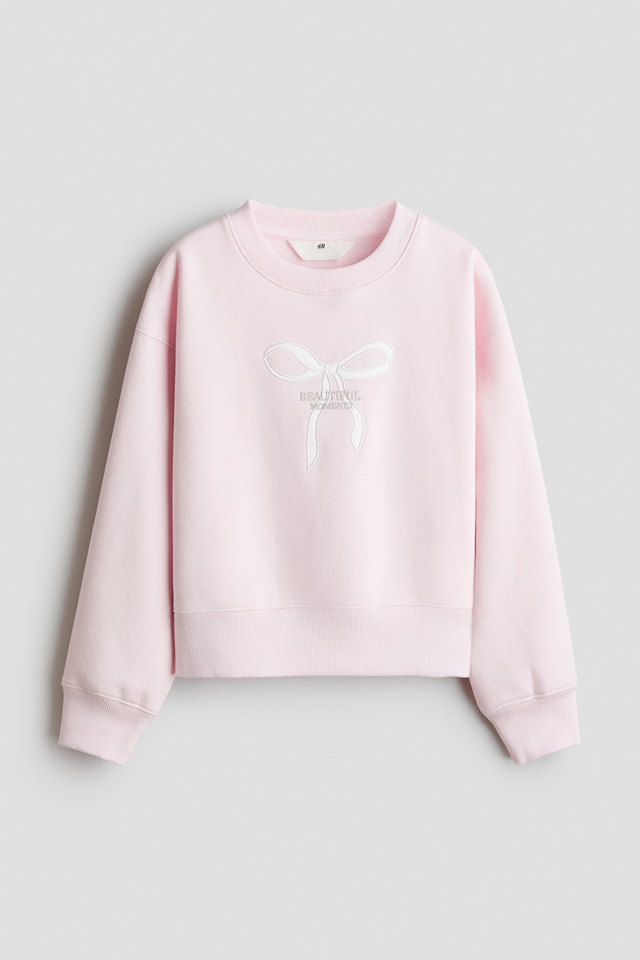 View larger image: A pink loose-fit sweatshirt is shown from the front, featuring a shimmering white bow graphic with "beautiful moments" text on the front, a round rib-trimmed neckline, dropped shoulders, ribbed cuffs, and a ribbed hem.