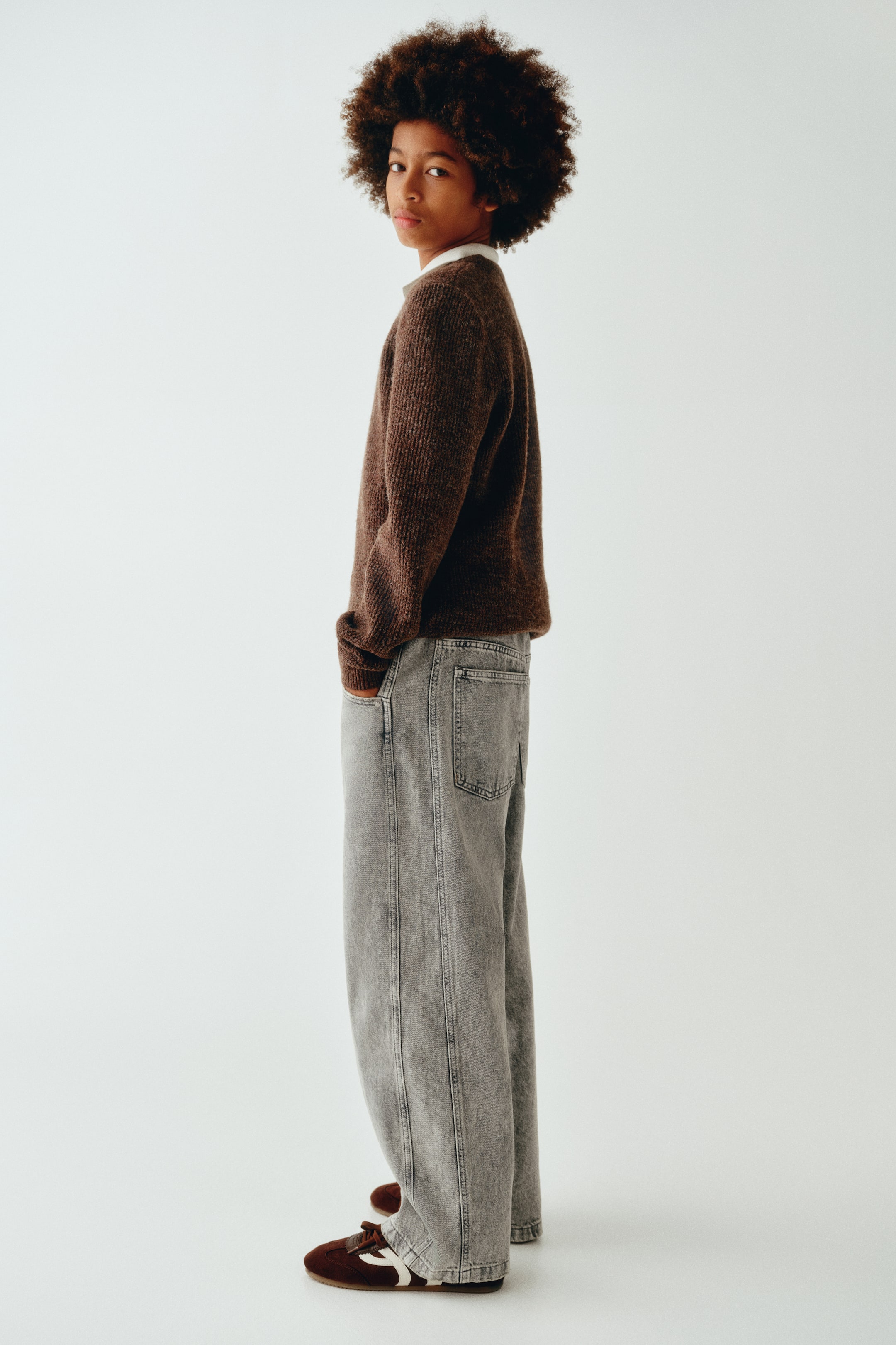 View larger image: Grey denim jeans in a loose fit are shown from a side-profile view, showcasing their relaxed, wide-leg silhouette, dropped crotch, and classic back pocket. The jeans are styled with a brown knit jumper and dark trainers, emphasising their casual drape.