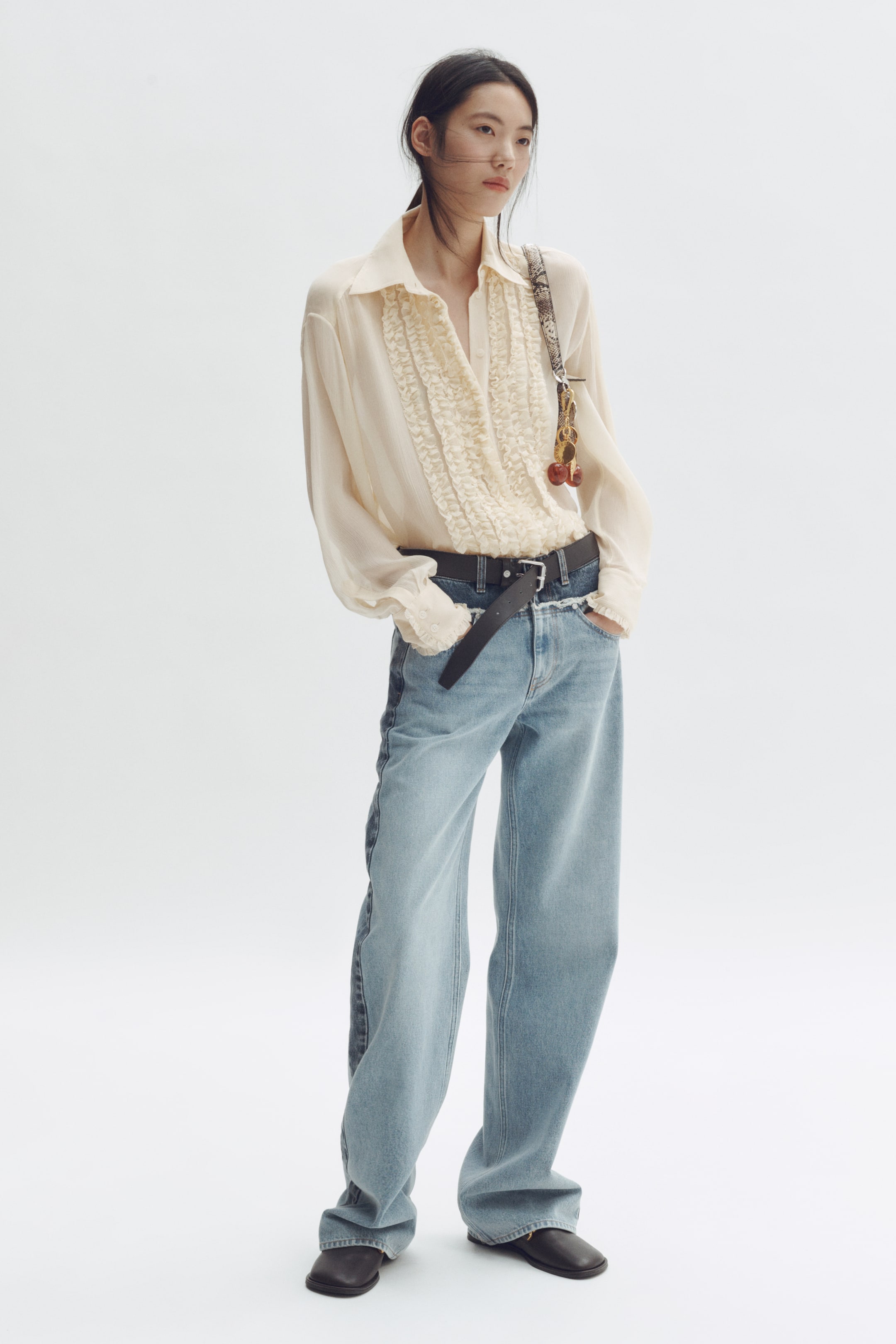 Größeres Bild anzeigen: Eine cremefarbene, transparente Bluse mit einer Rüschenleiste und gerafften Manschetten wird in eine hell gewaschene, weit geschnittene Jeans gesteckt getragen. Die Jeans präsentieren kontrastierende, dunkle Denim-Seitenpaneele und einen rohen Saum, gesichert durch einen schwarzen Gürtel.