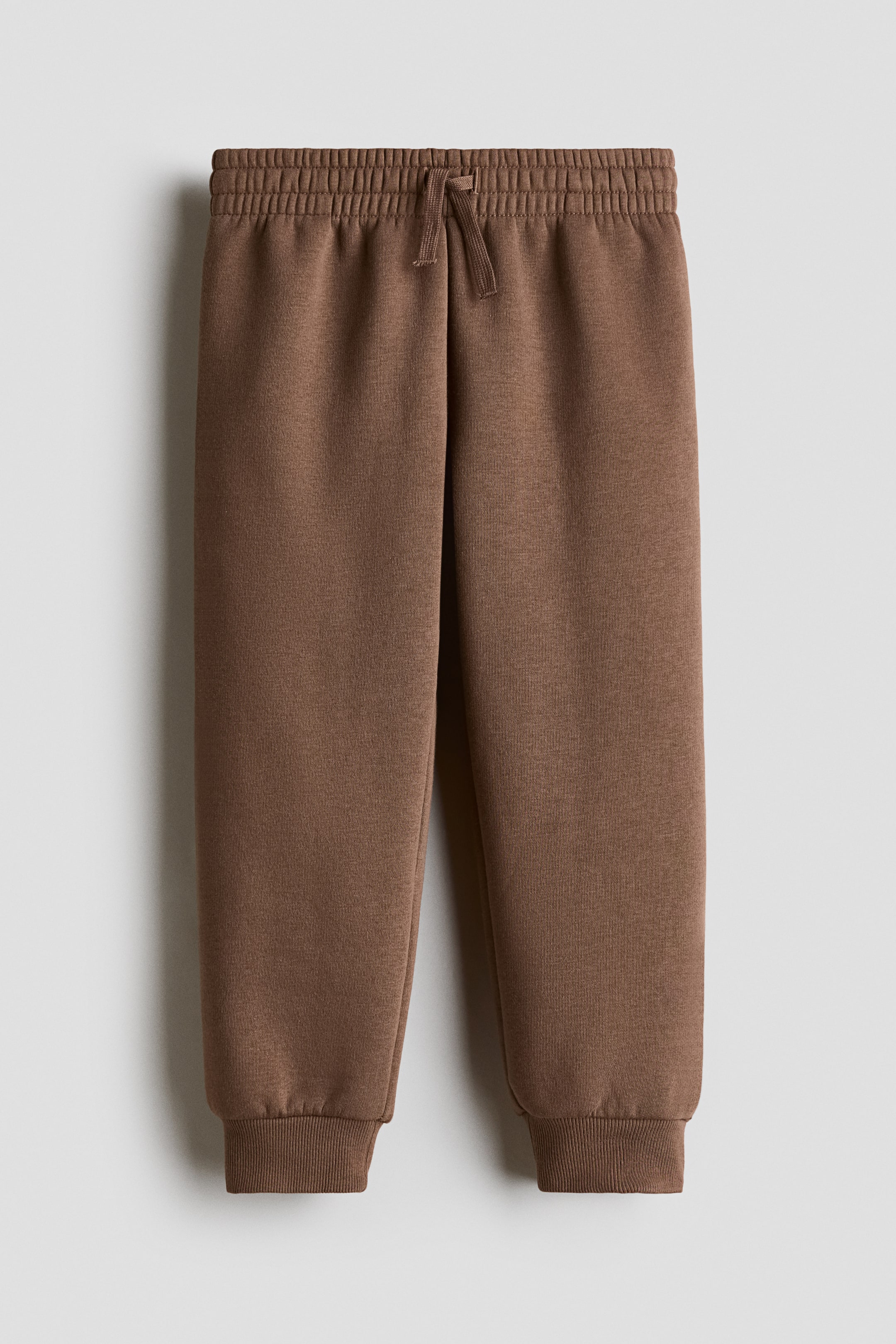 Agrandir l'image: Un pantalon de jogging marron est présenté de face, avec une taille élastiquée et un cordon de serrage noué. Le pantalon présente une texture subtile en tissu molletonné et se termine par des chevilles côtelées.