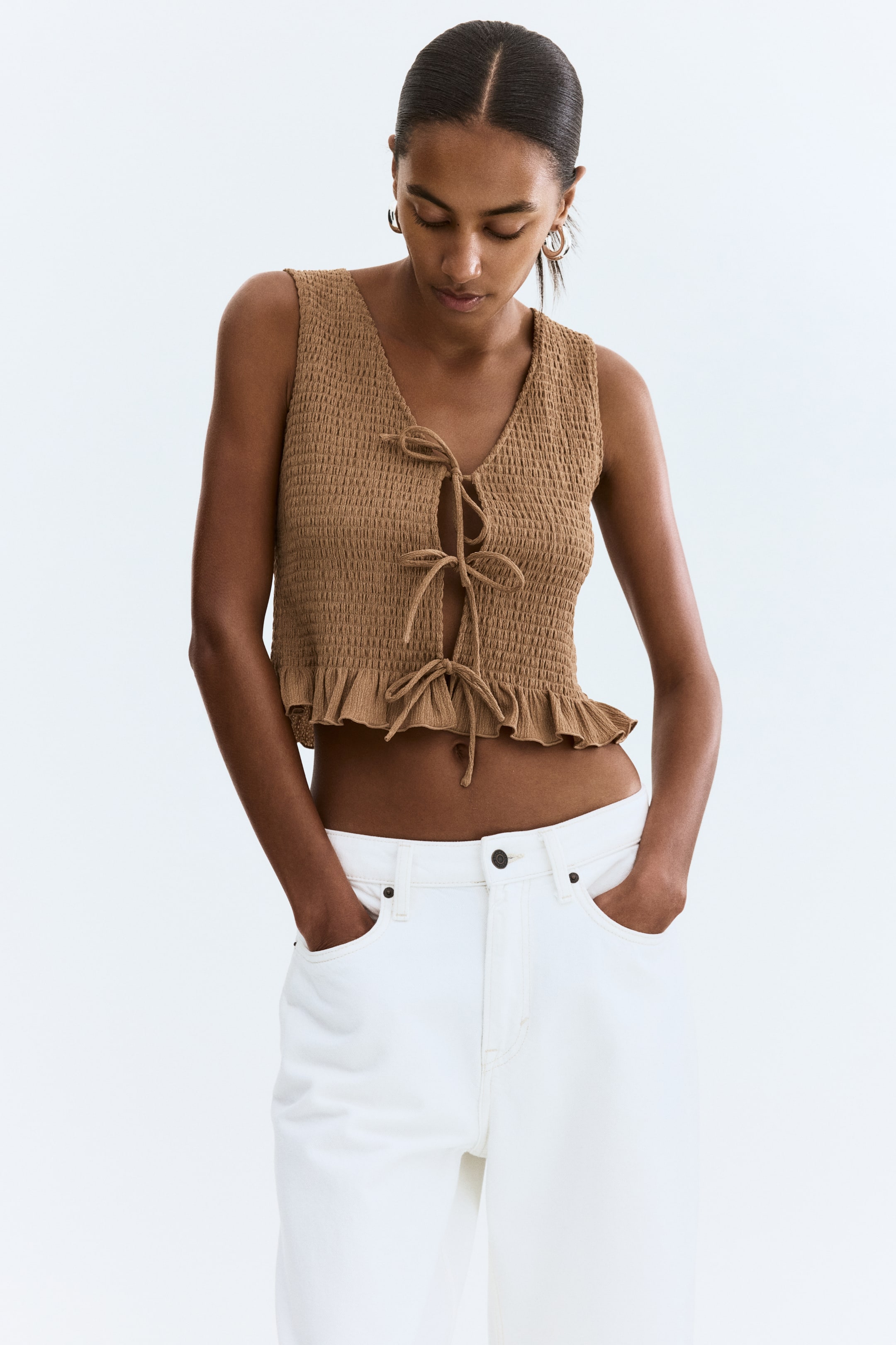 Xem ảnh lớn hơn: Áo không tay màu nâu có kết cấu nhăn, cổ chữ V, phía trước có dây buộc với nhiều dây thắt và gấu áo xếp nếp. Nó được kết hợp với quần trắng có túi trước và cài nút.