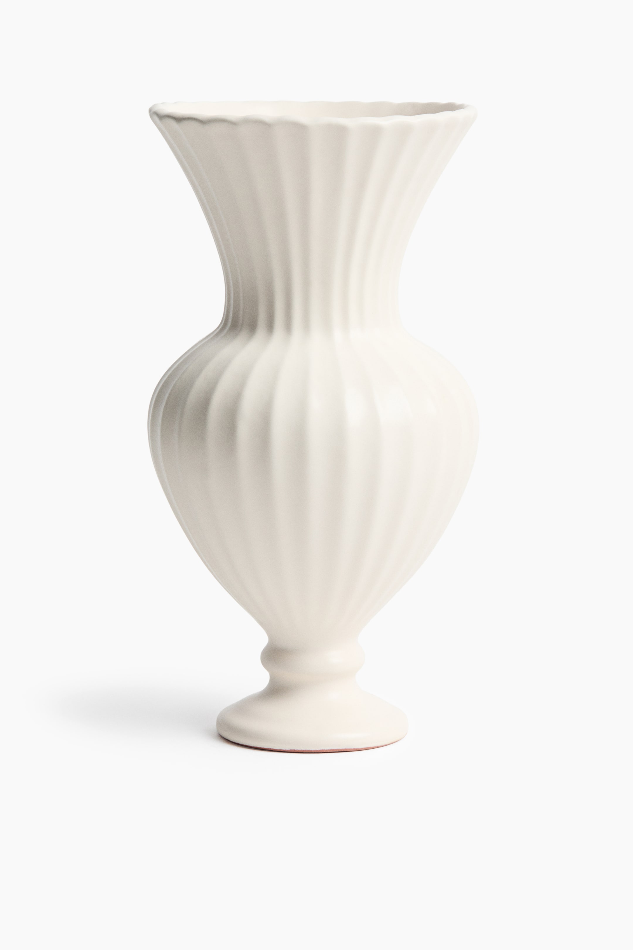 View larger image: A rounded, fluted terracotta vase stands upright, showcasing an off-white semi-matte glaze with a sliver of natural terracotta visible at the bottom rim. The vase tapers from a wide, flared rim with a scalloped edge into a narrow neck, expanding into a bulbous body with vertical fluting, and rests on a sculptural pedestal foot.
