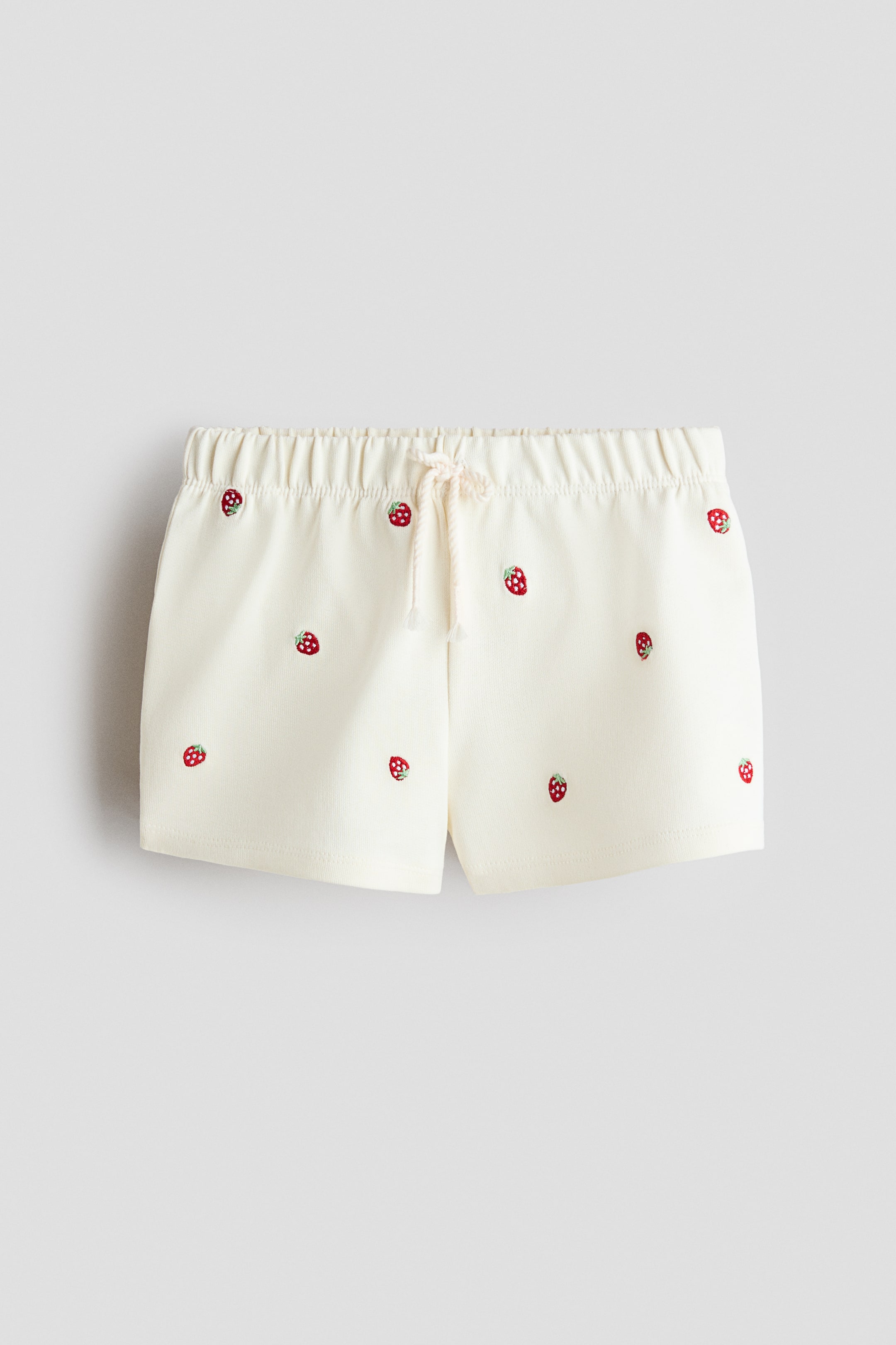 View larger image: The cream shorts feature an elastic, ruched waistband with an off-white drawstring tie. Small red strawberry embroideries are scattered across the front of the shorts.