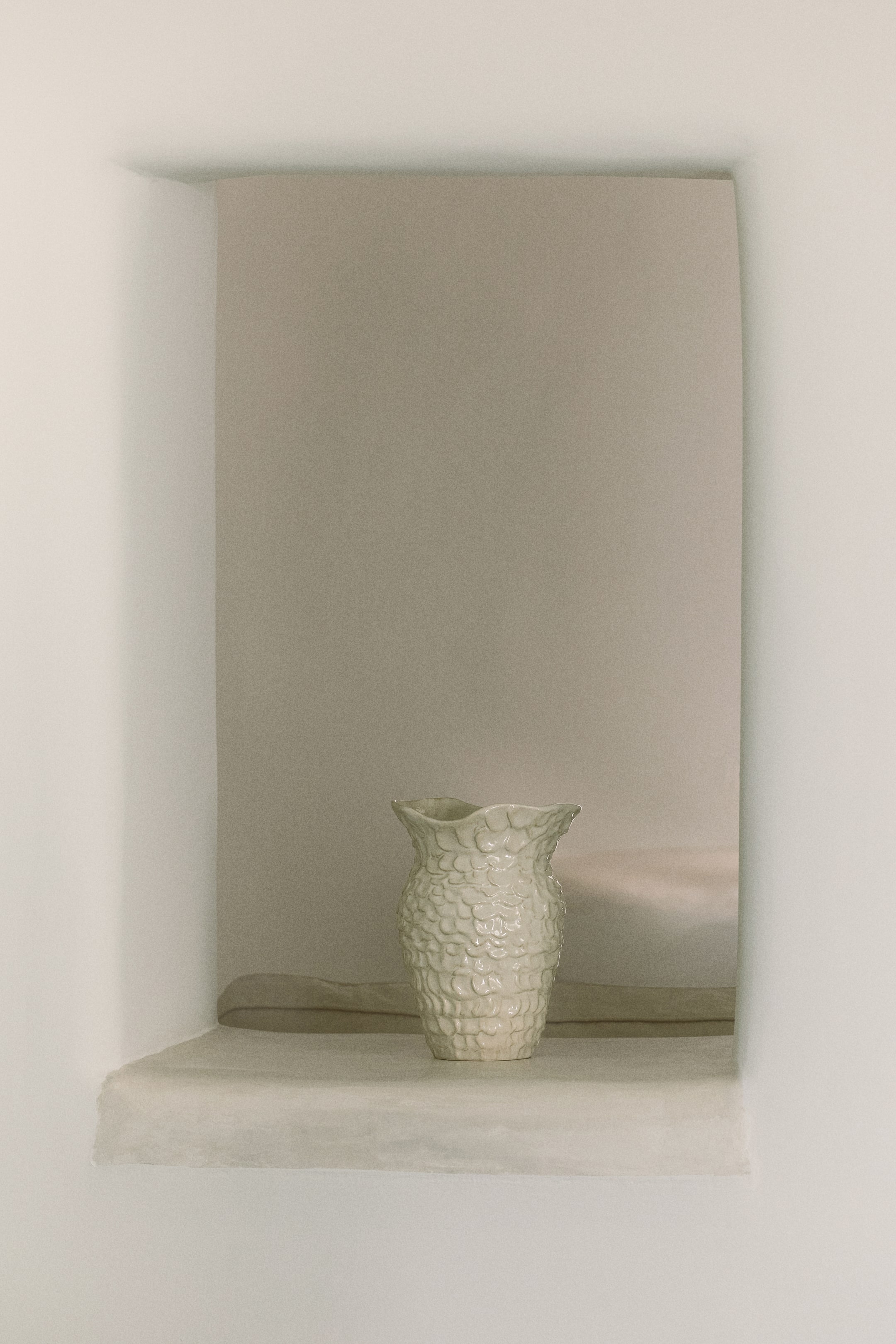 拡大画像を表示: 淡い色のセラミック製の花瓶は、その胴体に鱗のような質感のある模様が施され、波状のフルート状の縁を持っています。やや前方に向けて左側に配置されています。