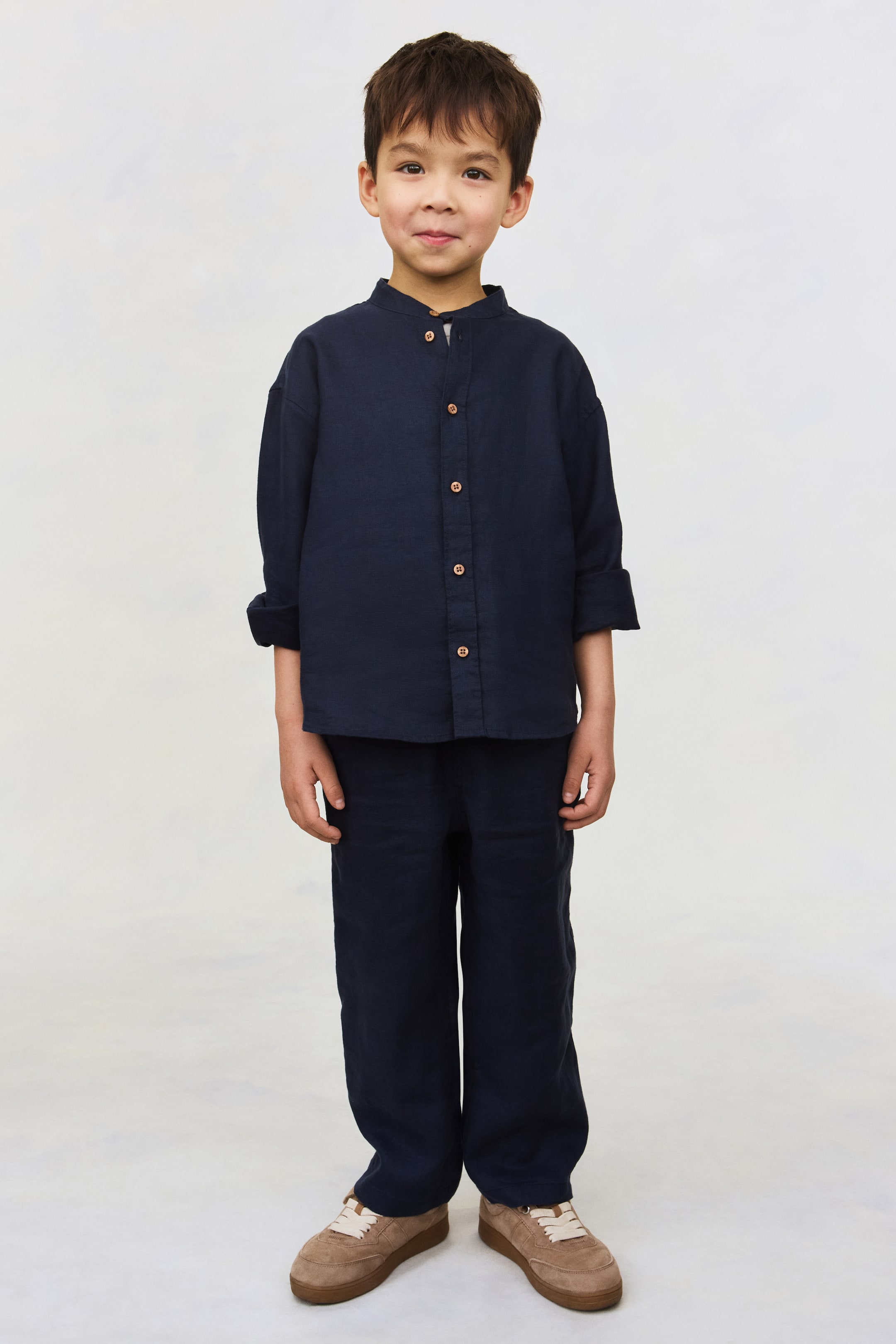 Agrandir l'image: Une chemise en tissu texturé bleu marine est portée, dotée d'un col mao, de boutons marron le long de la patte de boutonnage avant et de manches longues retroussées jusqu'aux coudes. Un pantalon assorti, ample et à jambe droite complète l'ensemble coordonné, présenté de face.