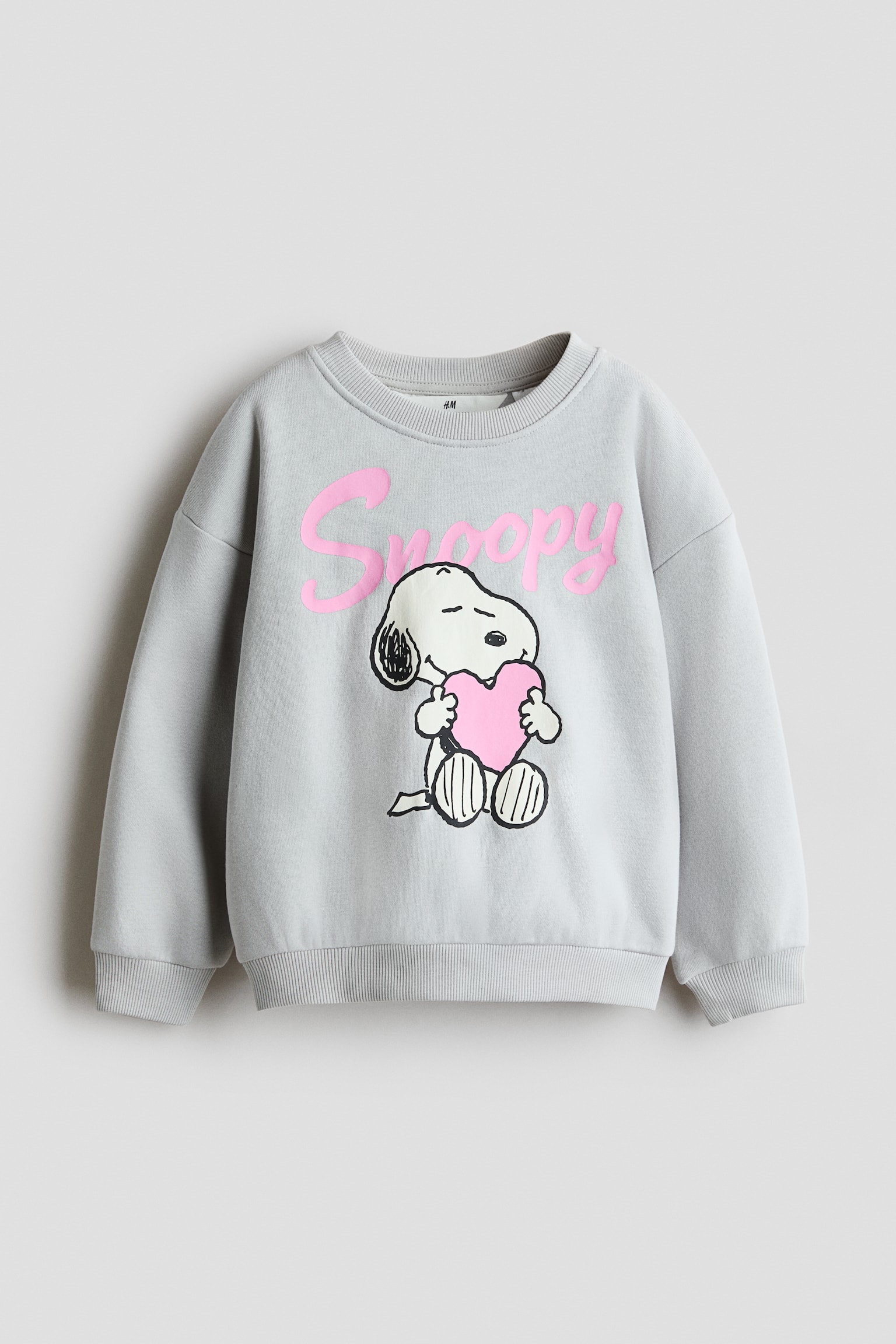 A light grey cotton-blend sweatshirt is shown from the front, featuring a printed graphic of the cartoon character Snoopy holding a pink heart with the name "Snoopy" in pink script above. The sweatshirt is styled with a round neckline, dropped shoulders, long sleeves, and ribbed trim at the collar, cuffs, and hem.