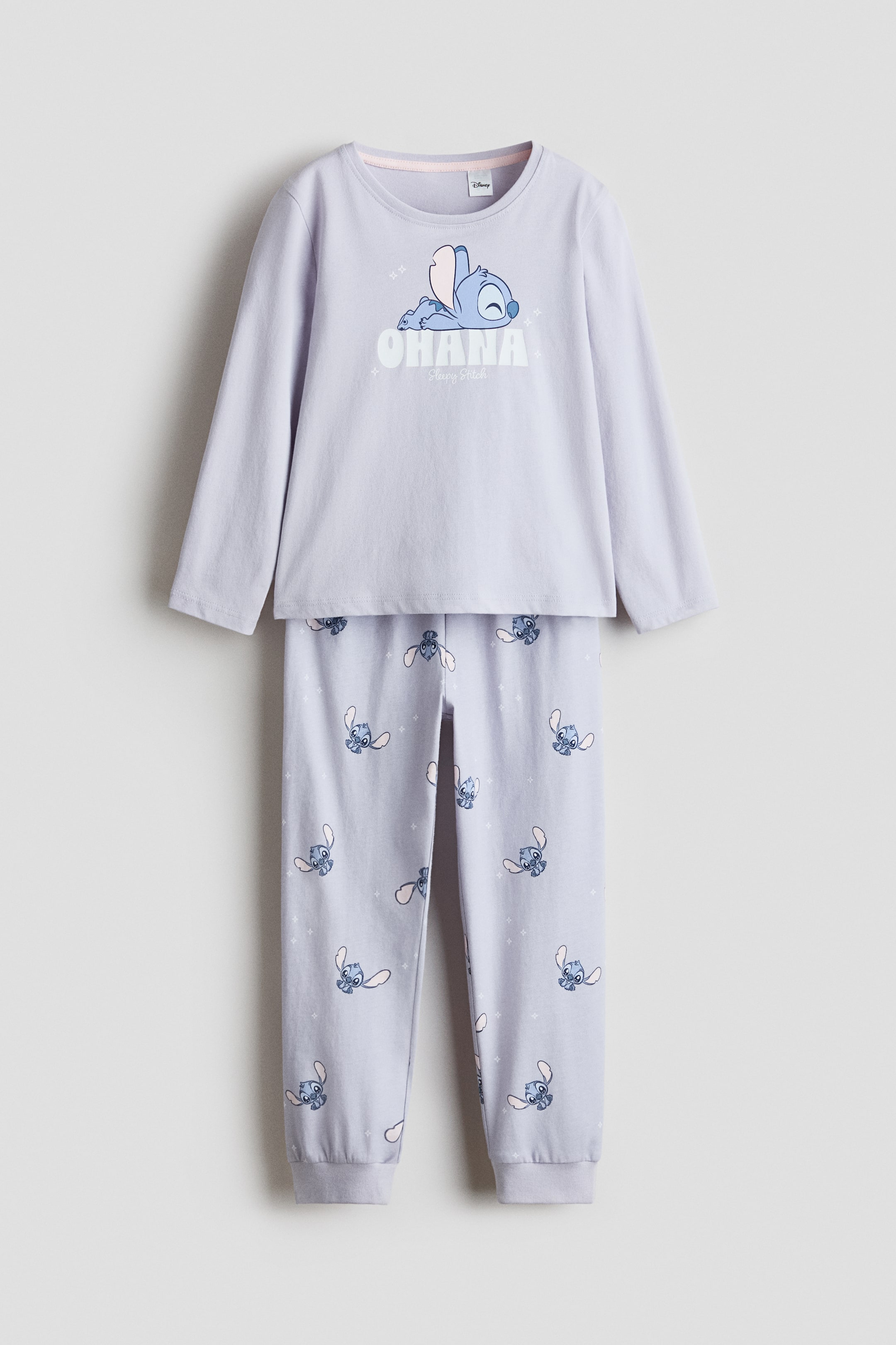 Näytä suurempi kuva: Vaalean violetti kaksiosainen pyjamasetti esitetään edestäpäin. Pitkähihaisessa yläosassa on grafiikka nukkuvasta Stitch-hahmosta ja teksti "OHANA Sleepy Stitch", ja yhteensopivissa pitkissä housuissa on toistuva kuvio pienemmistä Stitch-hahmoista ja valkoisista tähtikuvioista, jotka päättyvät resorillisiin nilkkamansetteihin.