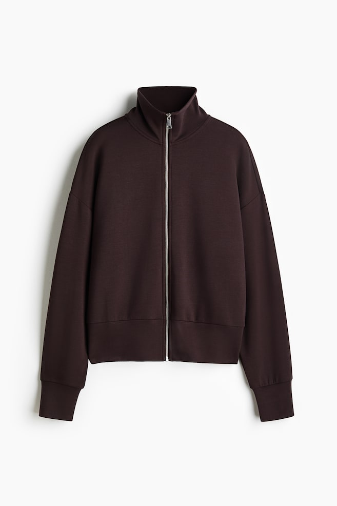 A loose-fit, dark brown track jacket is shown front-facing, featuring a stand collar and a silver zip extending down the front. It displays dropped shoulders, long sleeves with double-layered cuffs, and a double-layered hem. - 1