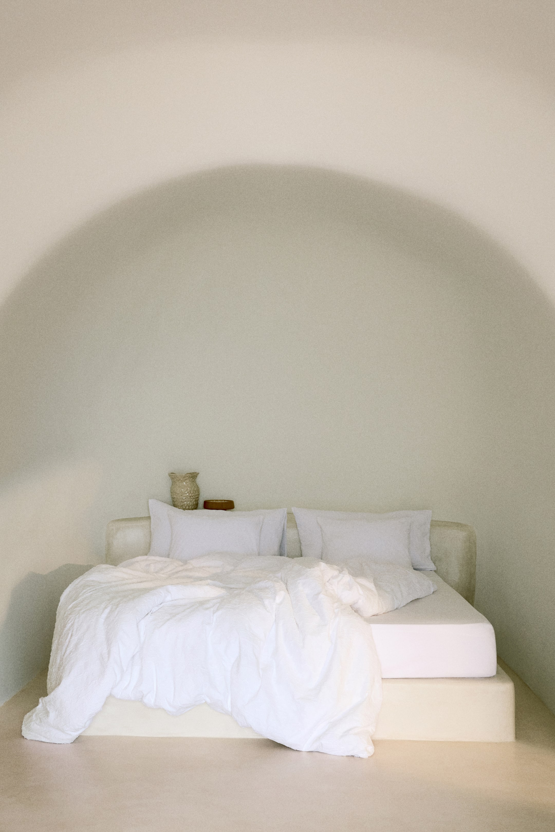 Voir une image plus grande: Un lit présente une base de plateforme solide de couleur claire. Un linge de lit blanc recouvre le matelas, incluant une couette blanche texturée drapée à l'avant et deux oreillers blancs reposant contre une tête de lit intégrée de couleur claire.