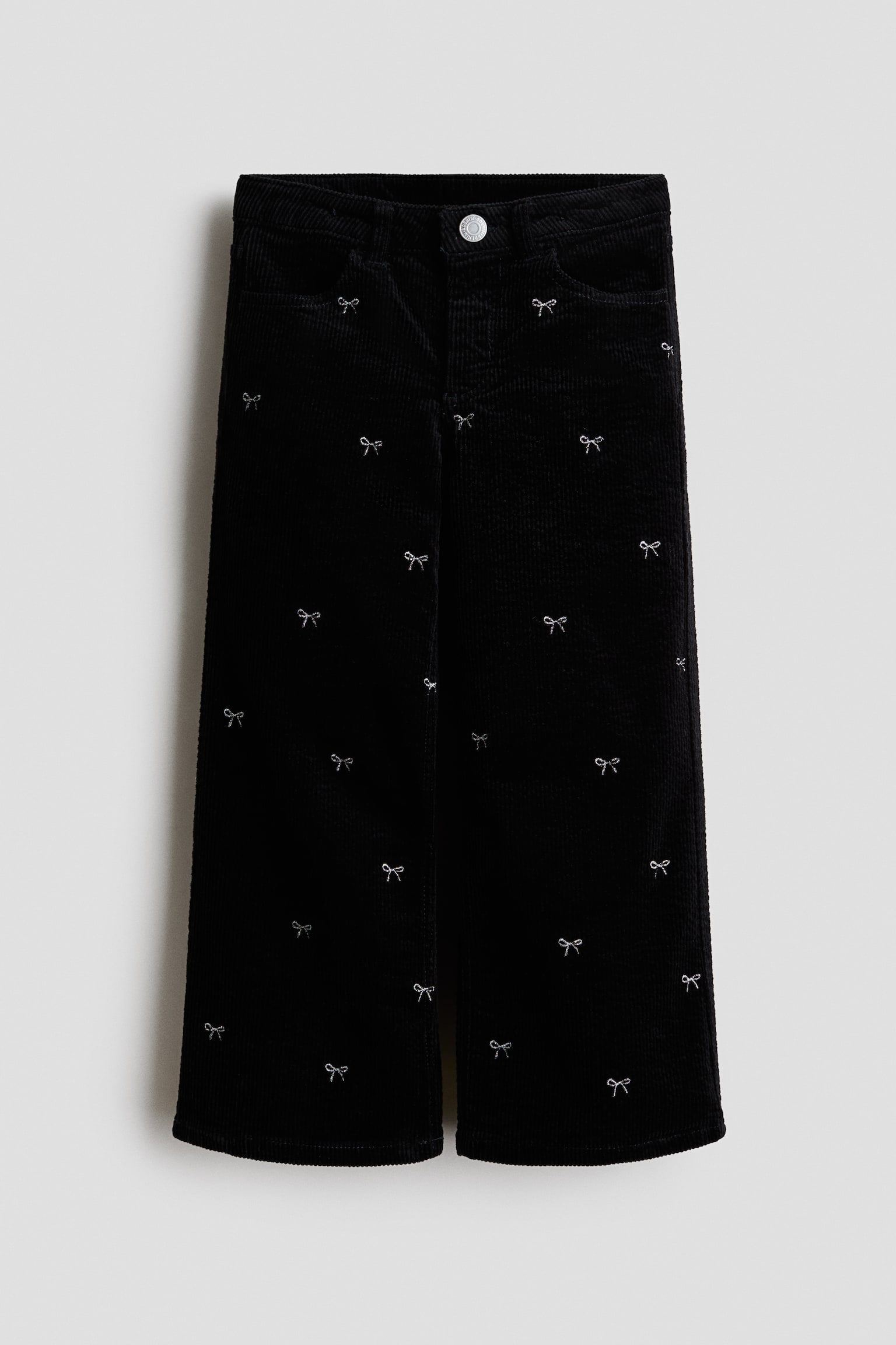 A pair of black corduroy trousers is shown from the front, featuring a straight-leg silhouette adorned with an all-over pattern of small embroidered silver bows. The trousers feature a classic waistband with a silver-tone button closure and front pockets.