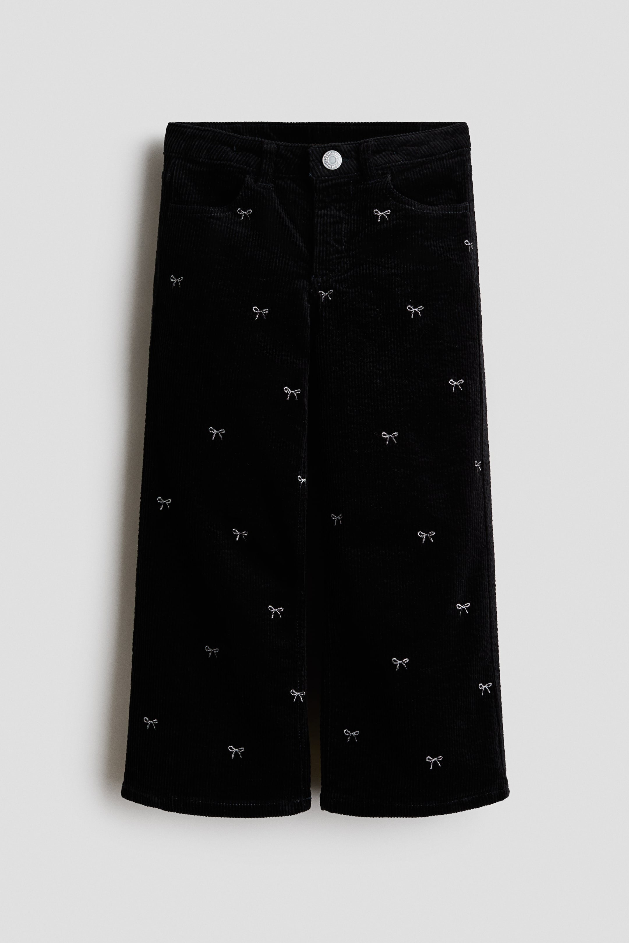 Voir une image plus grande: Une paire de pantalons en velours côtelé noir est présentée de face, mettant en valeur une silhouette à jambe droite ornée d’un motif intégral de petits nœuds argentés brodés. Les pantalons sont dotés d’une taille classique avec une fermeture à bouton de couleur argentée et de poches avant.