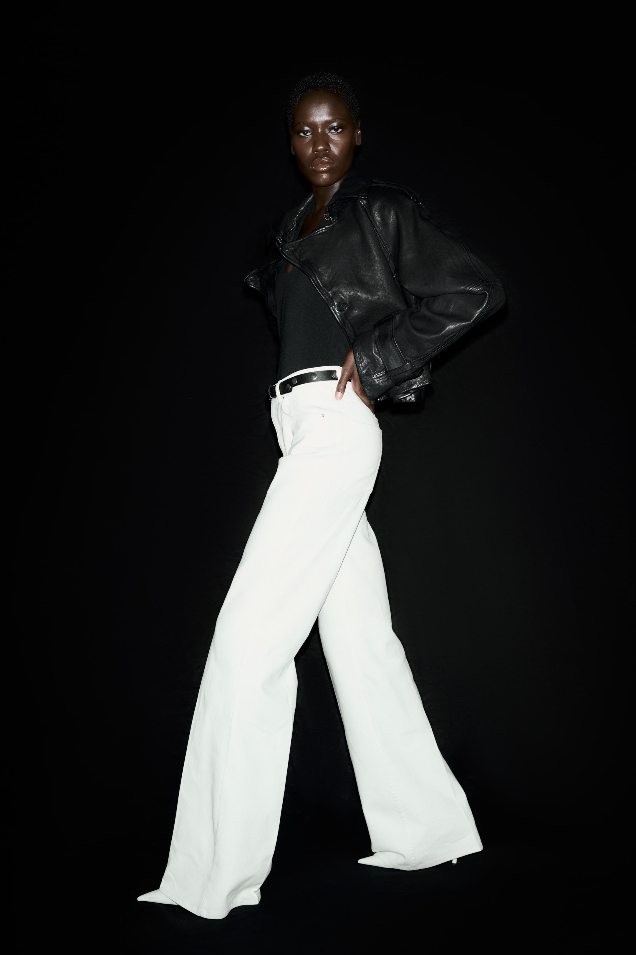 Voir une image plus grande: Le mannequin porte un pantalon blanc ajusté. Ils présentent une taille haute avec une ceinture cloutée noire, des poches avant ouvertes et des jambes évasées avec des plis à l'avant. Le mannequin pose avec une main sur la hanche.