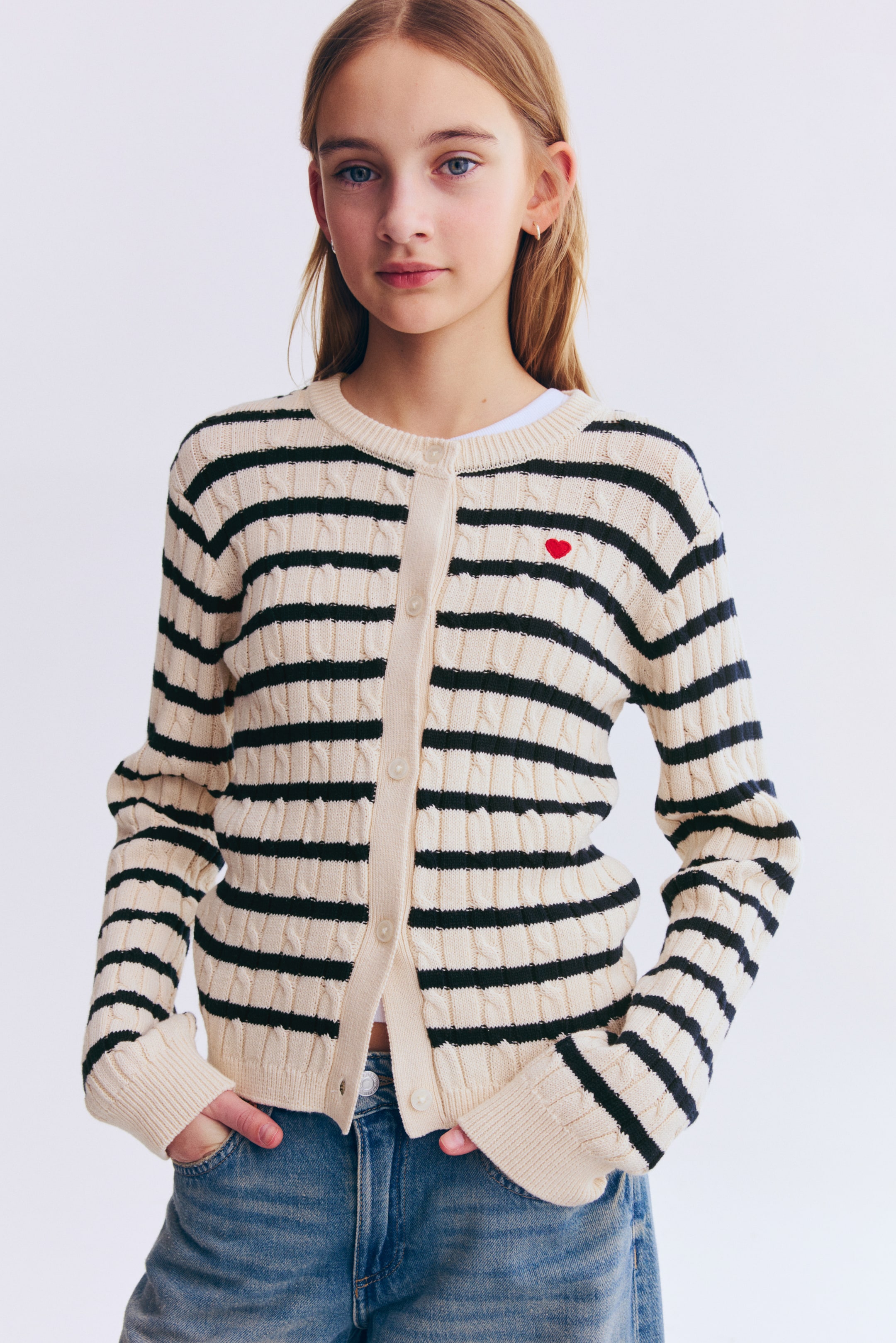 View larger image: A cream and black striped cable-knit cardigan featuring horizontal black stripes, a crew neck, and a button-front closure with cream buttons. A small red embroidered heart appears on the left chest, and the long sleeves end in ribbed cuffs.