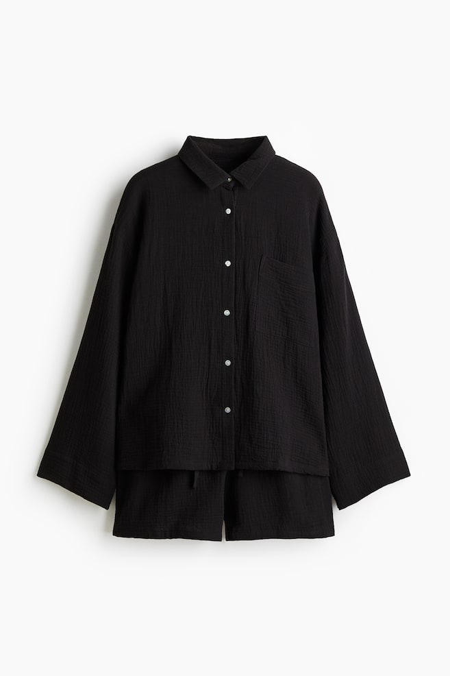 A black pyjama set is presented from the front, consisting of a long-sleeved shirt and matching shorts in textured cotton muslin. The shirt is featuring a collar, light-colored buttons down the front, and a patch chest pocket. The shorts are designed with an elasticated drawstring waist. - 1