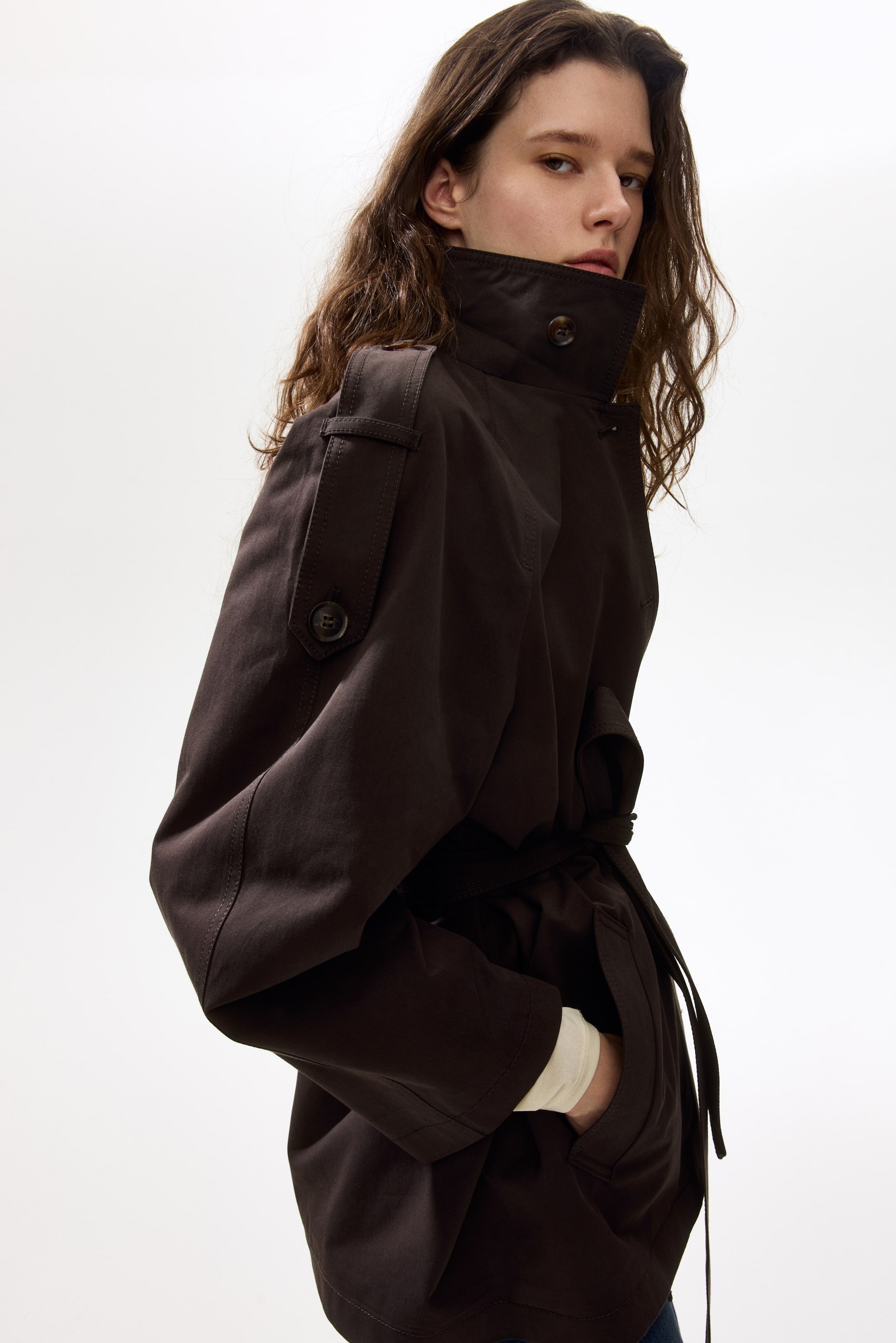 Agrandir l'image: Une veste marron foncé est présentée sous un angle de côté avant, avec un col montant, des épaulettes à bouton et une ceinture en tissu assorti cintrant la taille. Une main repose dans une poche latérale, révélant une manche de couleur claire qui dépasse du poignet de la veste.