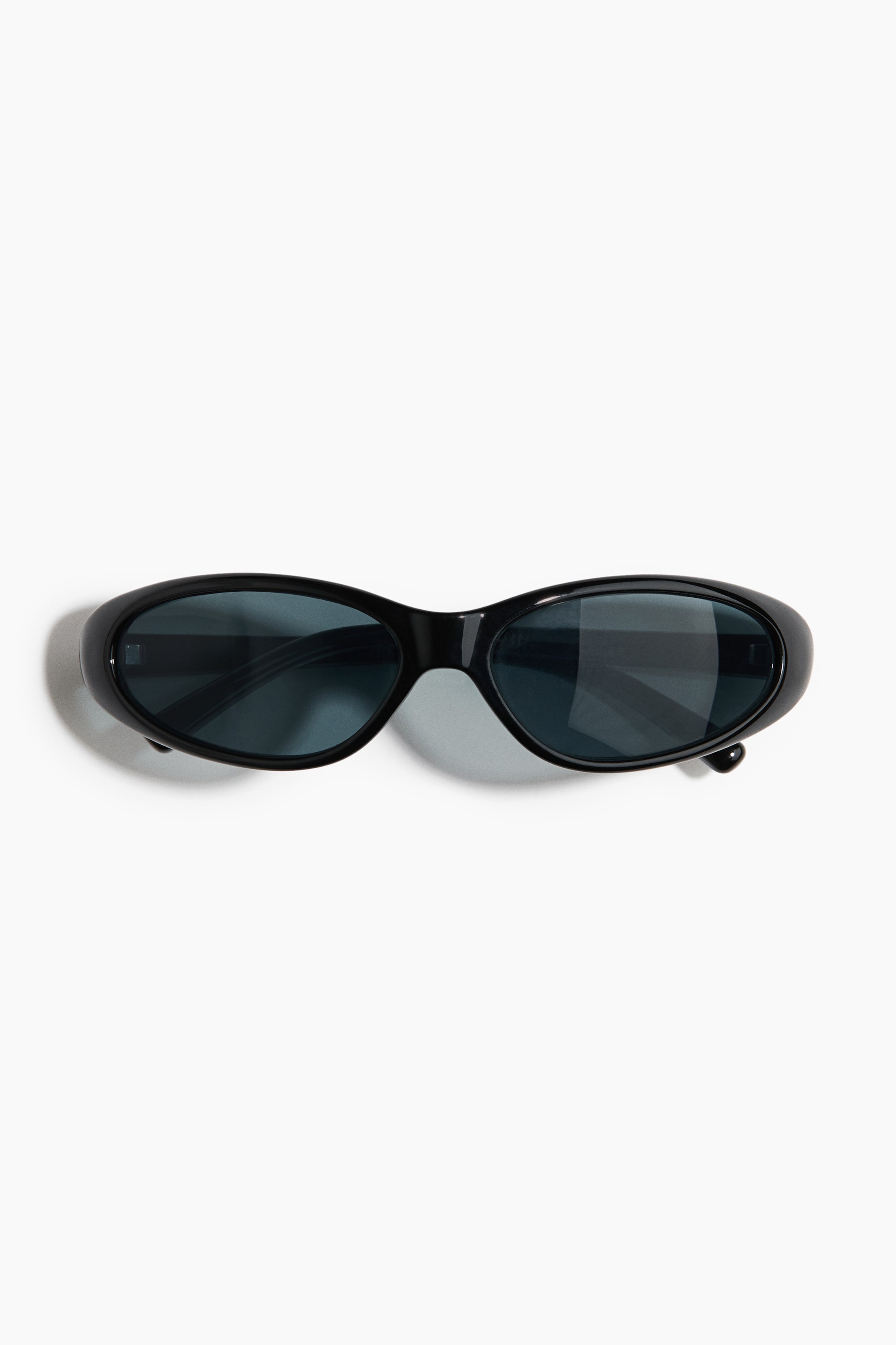 Größeres Bild ansehen: Die Sonnenbrille präsentiert sich mit einem eleganten, schwarzen Gestell und elliptischen, dunkel getönten Gläsern, die flach liegen und nach vorne gerichtet sind.