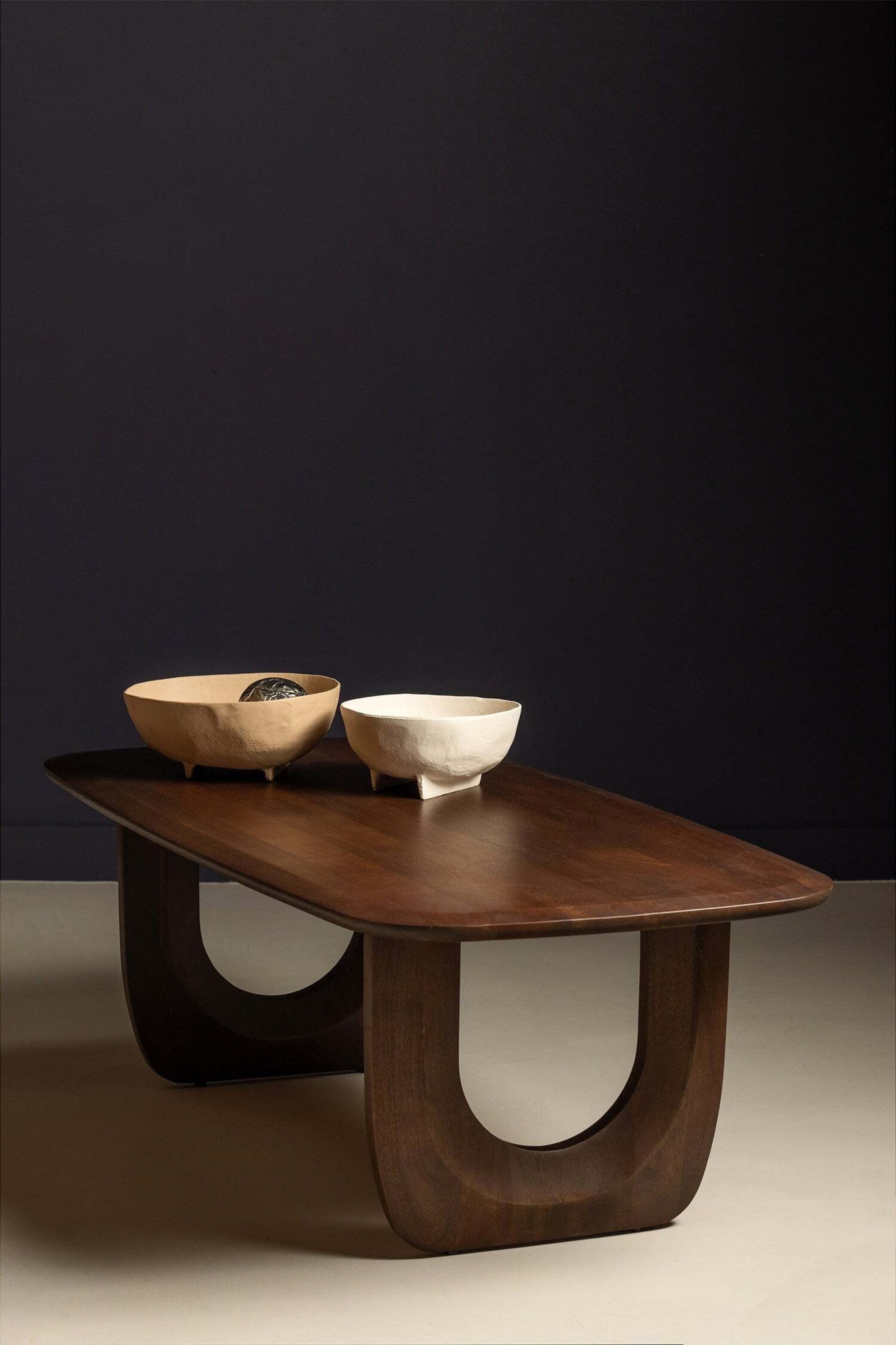 Agrandir l'image: Une table basse en bois foncé présente un plateau allongé et arrondi. Elle repose sur deux pieds épais en bois en forme de U.