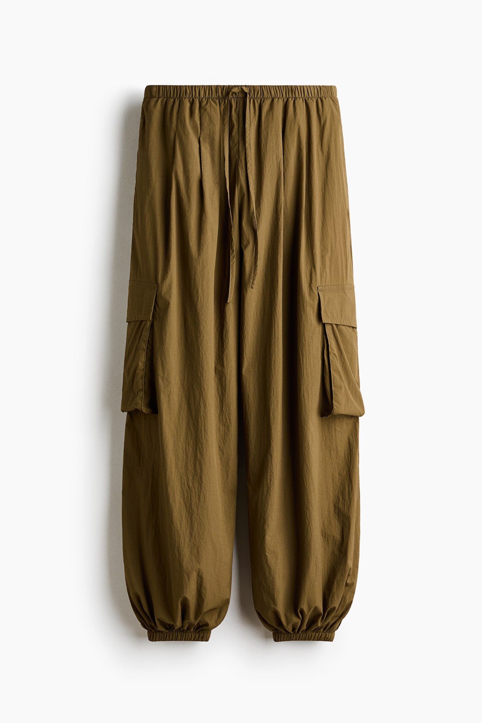 Estos pantalones de nailon de gran tamaño, en color marrón oliva, cuelgan vistos de frente, mostrando una cintura elástica con cordón y voluminosas piernas tipo globo con pinzas en la parte superior. El diseño presenta grandes bolsillos cargo tipo fuelle con solapas a los costados y se estrecha en puños fruncidos con elástico.