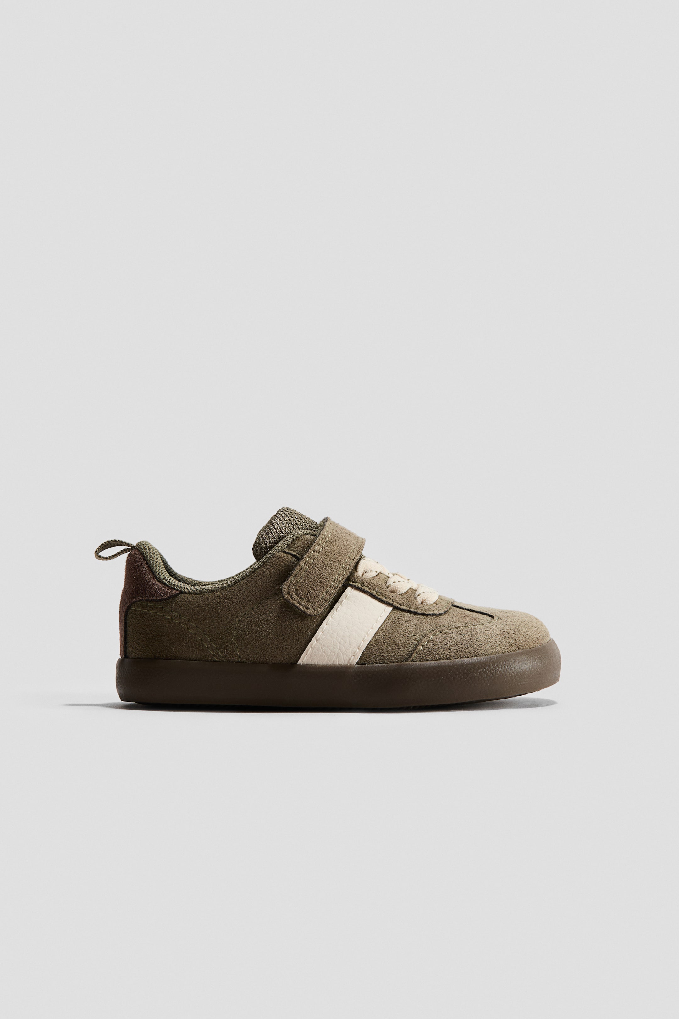 View larger image: Barefoot trainer. It features an olive green suede-like upper with a single hook-and-loop strap closure, elasticated cream laces, and a prominent cream stripe on the side. The trainer showcases a mesh tongue and collar, a dark brown heel accent, a small pull tab at the heel, and a thin, flat dark brown sole.
