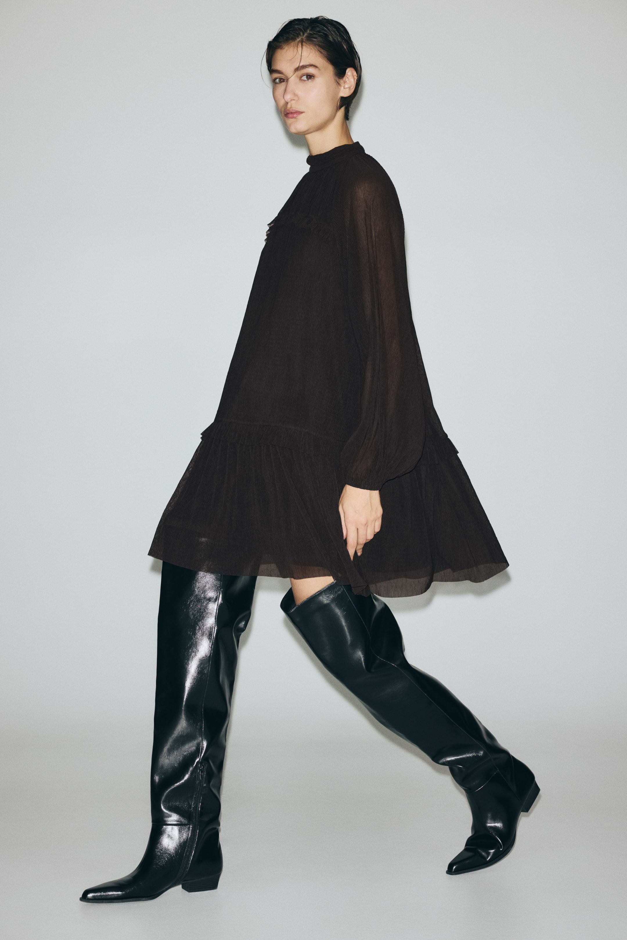 View larger image: A dark brown tiered dress features a high neckline with subtle pleating and long, sheer, voluminous sleeves gathered at the cuffs. The loose, flowing silhouette showcases a ruffled tier above the hem, appearing in motion from a three-quarter front view.