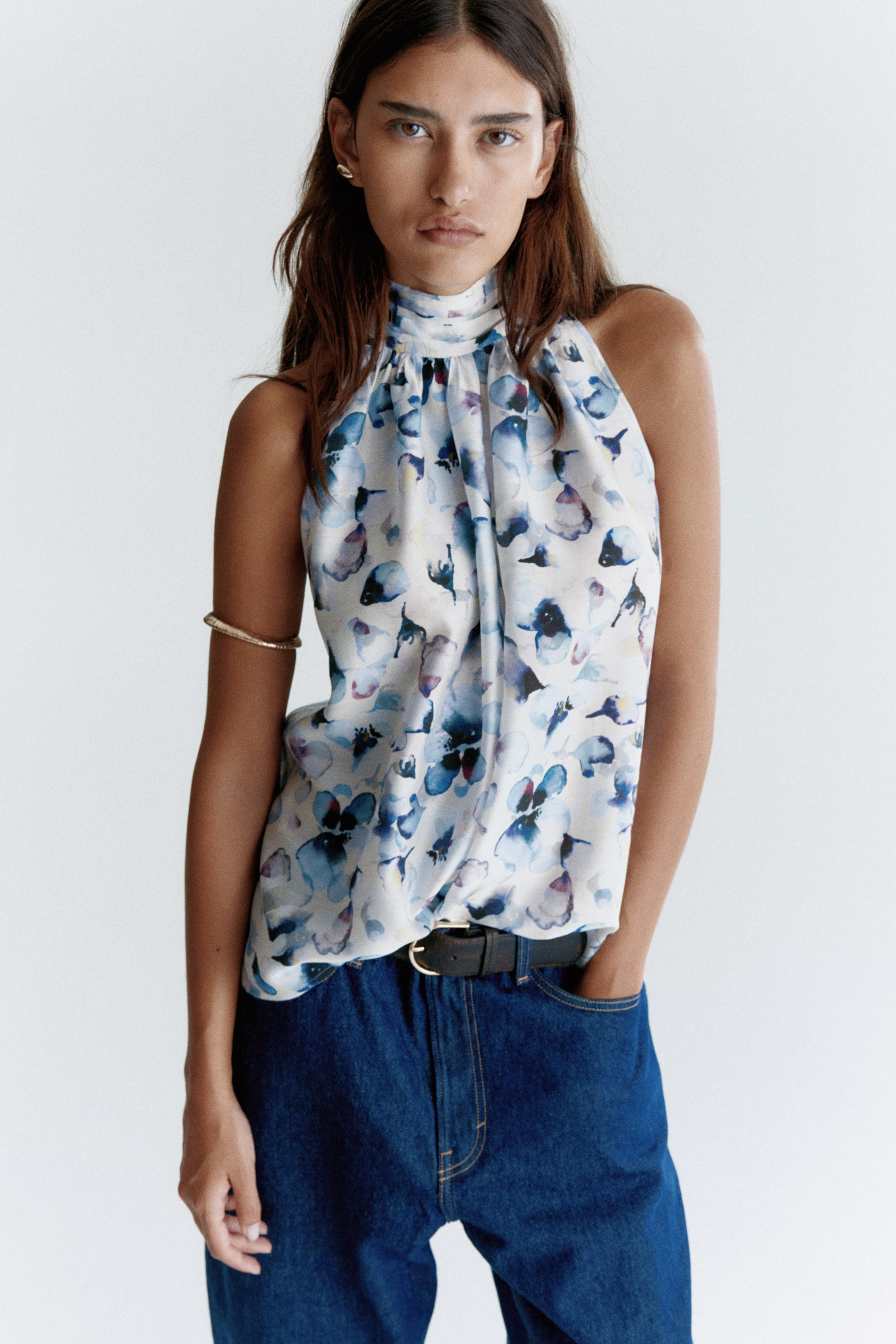 View larger image: A light-colored halter neck top, featuring a watercolor-style abstract floral pattern in shades of blue and purple, is being worn. It is designed with a high, gathered neckline and a sleeveless cut, currently tucked into dark denim jeans.