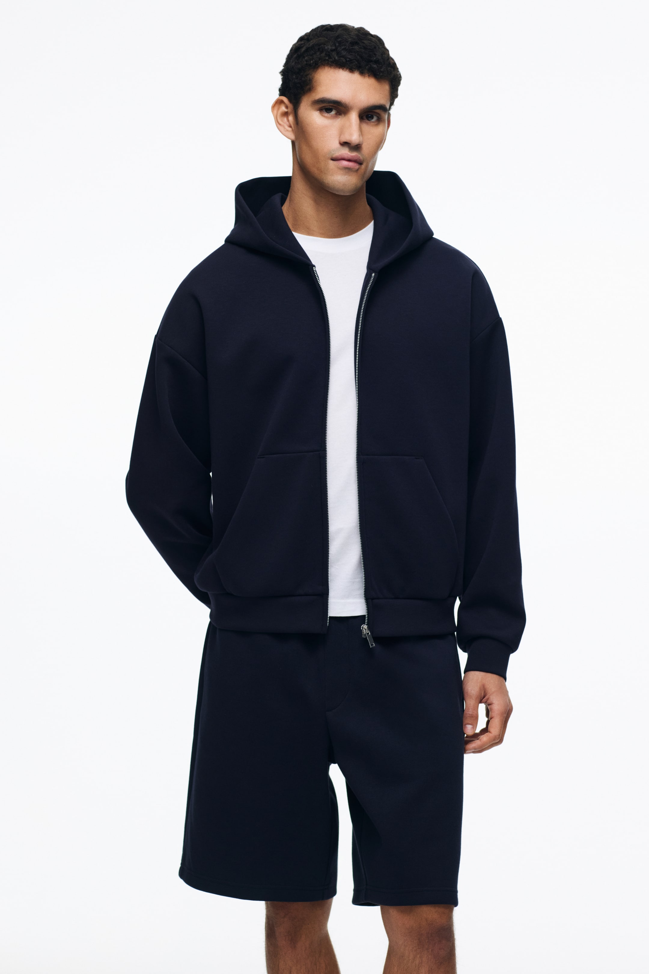 View larger image: Presenting a navy blue full zip hoodie paired with matching knee-length shorts, showcased from the front. The hoodie features a hood, dropped shoulders, ribbed cuffs and hem, and is worn open to reveal a white t-shirt underneath. The shorts are designed with a drawstring waist, offering a comfortable fit.