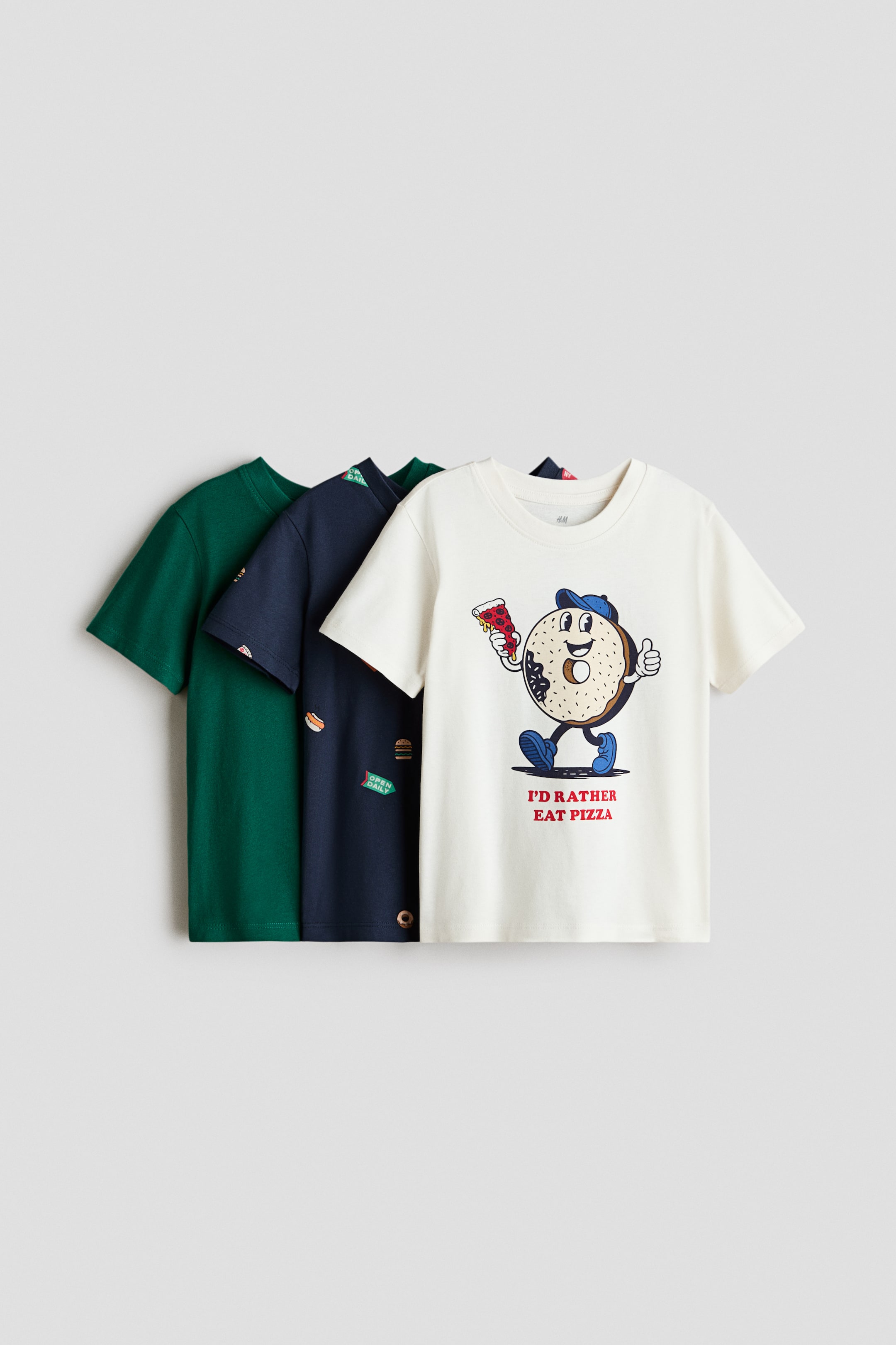 Grotere afbeelding bekijken: Drie klassieke T-shirts in katoenen jersey hebben ronde, geribbelde halslijnen en recht afgesneden zomen. Het voorste off-white T-shirt toont een gedrukt cartoon bagelkarakter dat een pizzapunt vasthoudt en tekst, terwijl het marineblauwe T-shirt daarachter een all-over print van fastfooditems toont, en het achterste donkergroene T-shirt een effen kleur heeft.