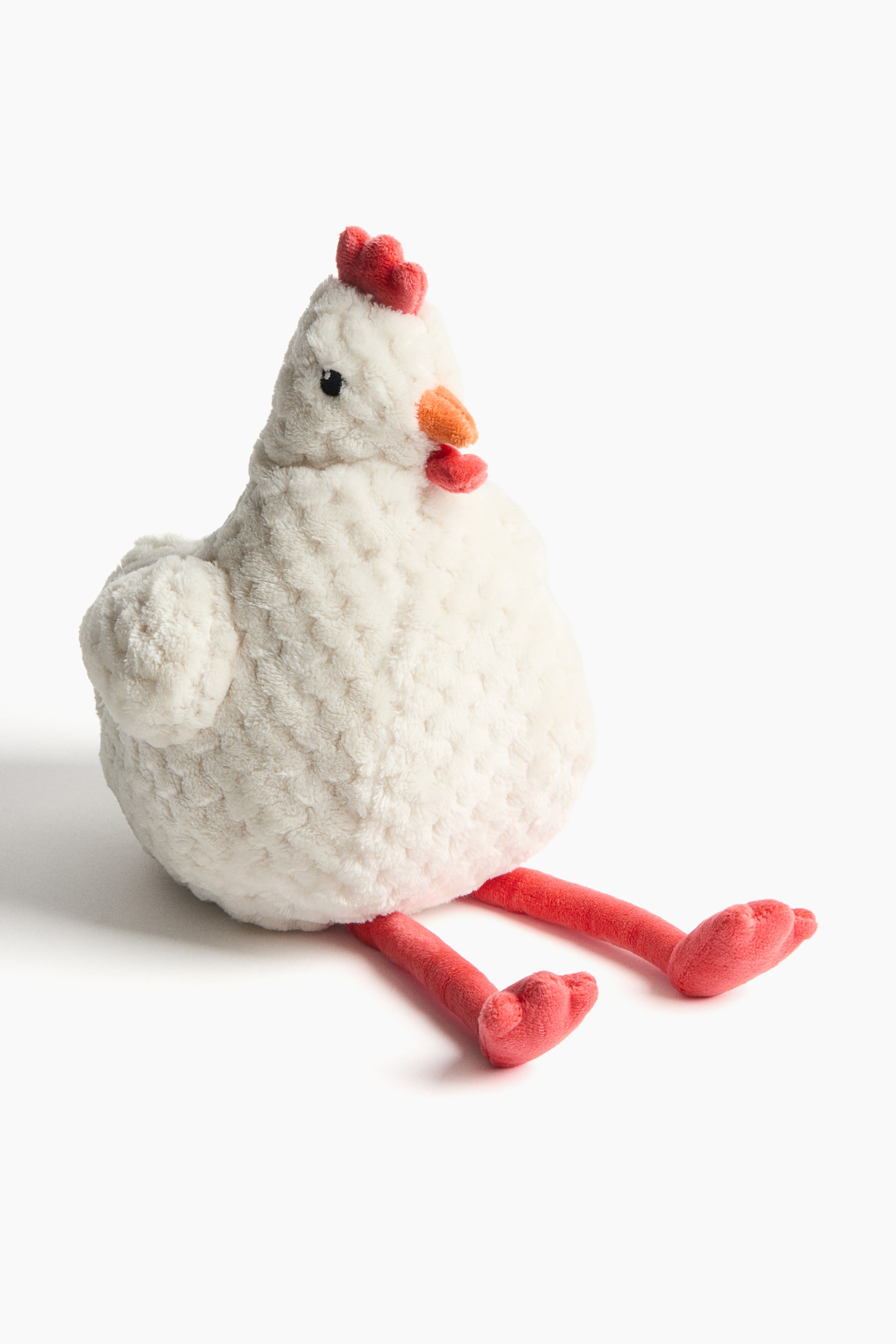 View larger image: A plush toy chicken is sitting, its fluffy white body turned slightly toward the front and side. It features a red comb, a red wattle, an orange beak, and small black eyes. Its long, slender red legs with matching feet extend forward.