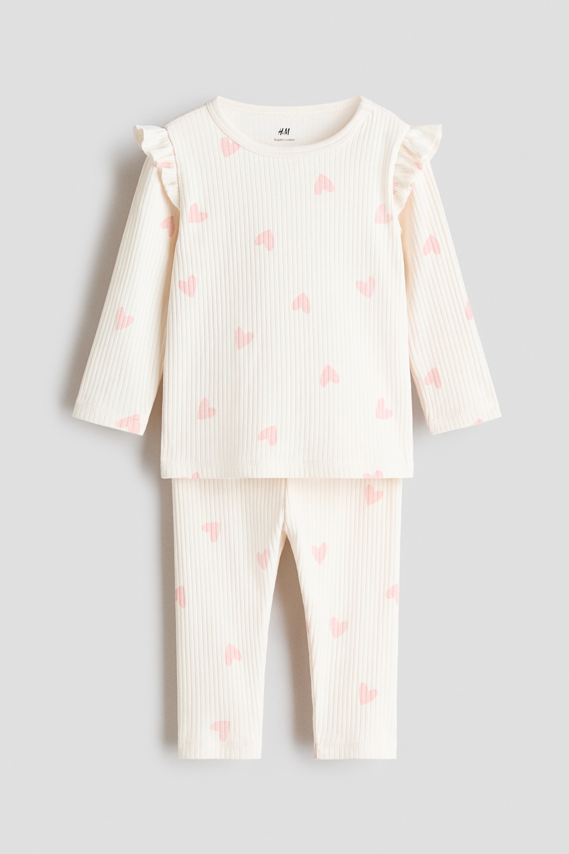 View larger image: A ribbed cotton set is displayed front-facing, comprising a cream long-sleeved top and matching full-length pants. The top shows a round neck and ruffled details on the shoulders, while both pieces feature a pattern of scattered pink hearts.