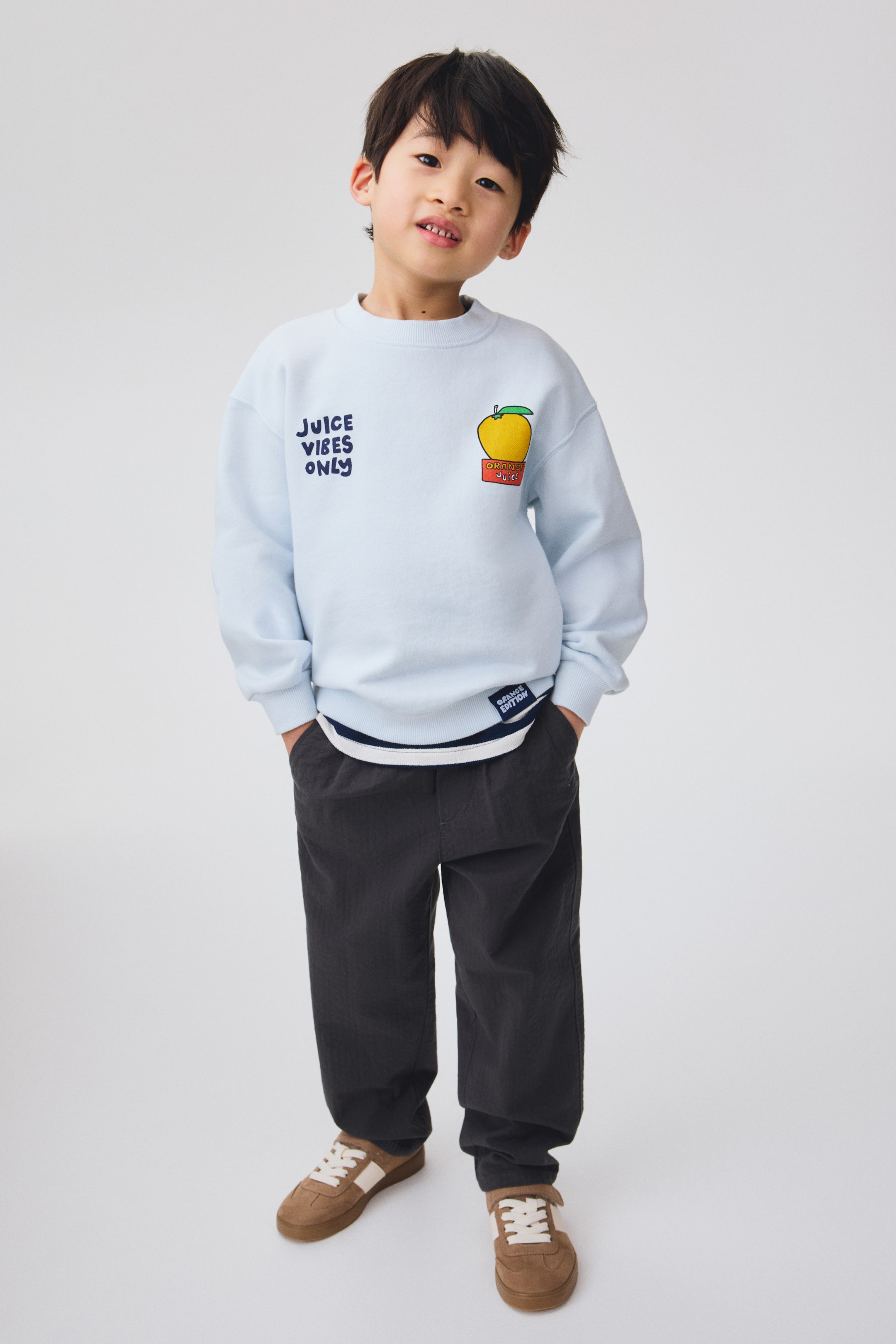 Büyük resmi görüntüle: Açık mavi bisiklet yaka bir sweatshirt, sol göğüs kısmında "JUICE VIBES ONLY" yazısı ve sağ göğüs kısmında "ORANGE JUICE" etiketli kırmızı bir kutu üzerinde sarı bir elma grafiği ile dikkat çekiyor. Önden görünüşlü bu sweatshirt, ribanalı manşetler ve etek ucu ile beyaz ve mavi çizgili bir gömlek katmanını etek ucunda görünür şekilde sergiliyor.