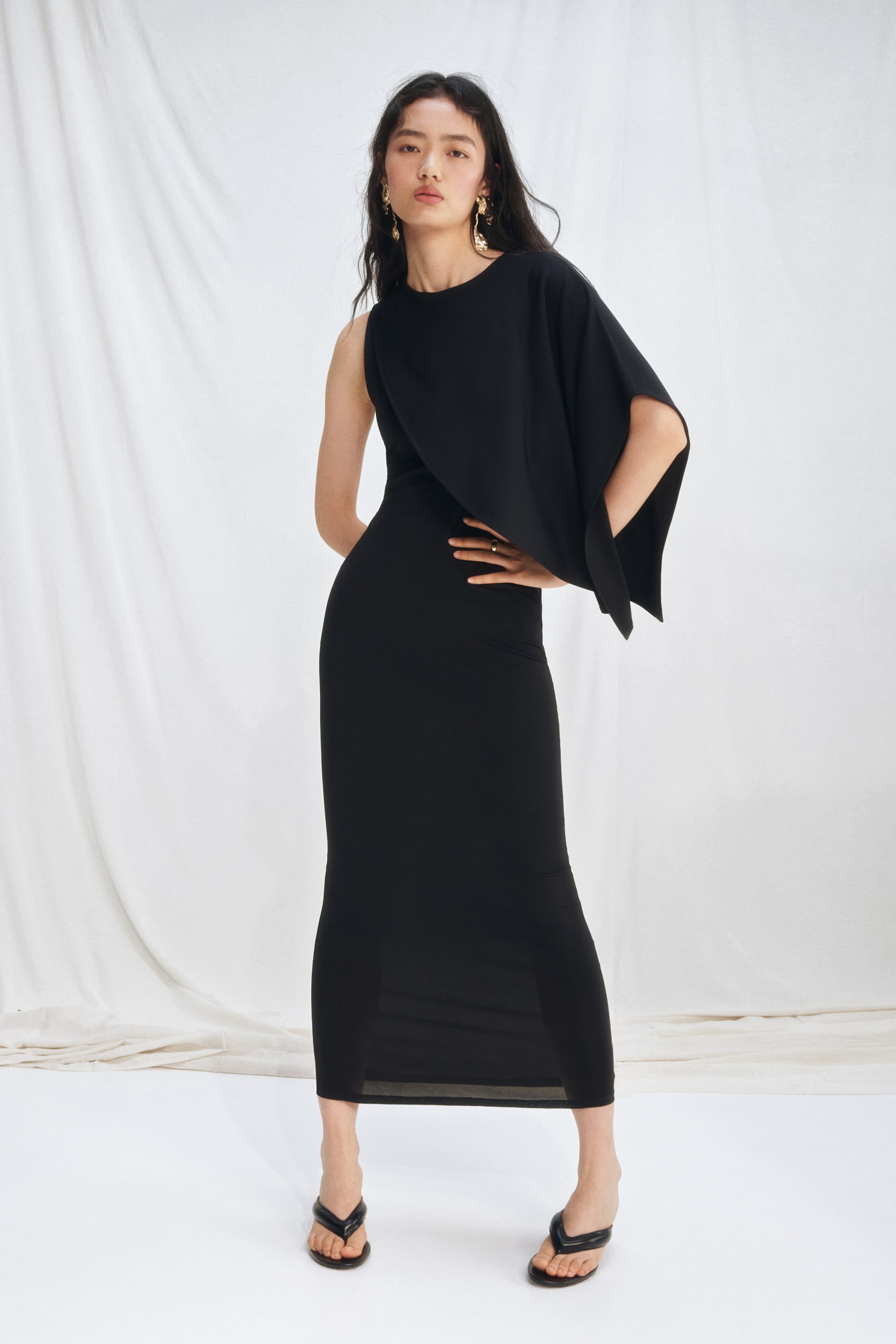 View larger image: A black maxi dress featuring an asymmetrical design, presenting one sleeveless shoulder and a wide, draped sleeve on the other. It showcases a rounded neckline and a form-fitting silhouette extending to the ankles.