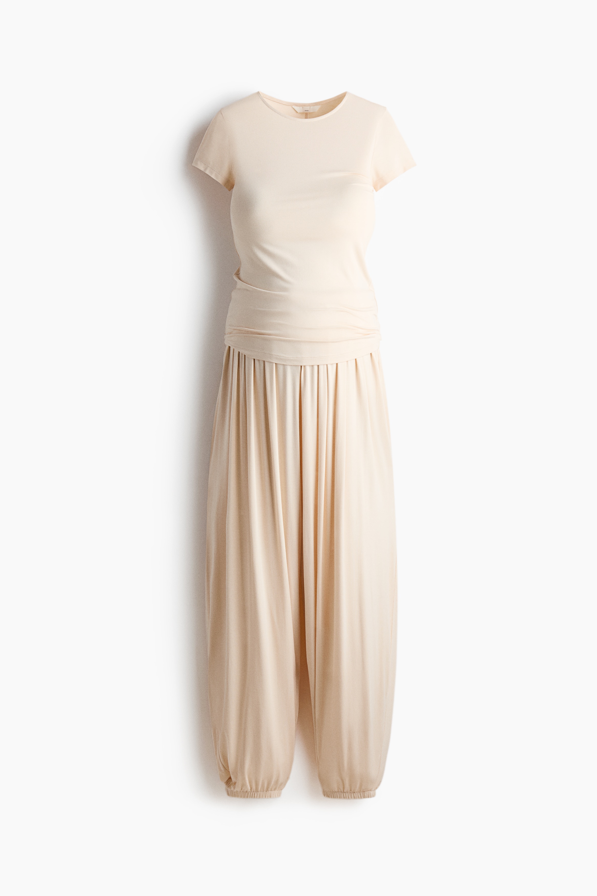 Een crÃ¨mekleurige T-shirt- en broekenset in stretch viscose jersey wordt gedragen door het model, met een aansluitend T-shirt met ronde hals en een losse, soepelvallende broek met elastische taille en trekkoord. Een bijpassende geribde gebreide trui is nonchalant over de schouders van het model gedrapeerd als gelaagd kledingstuk.