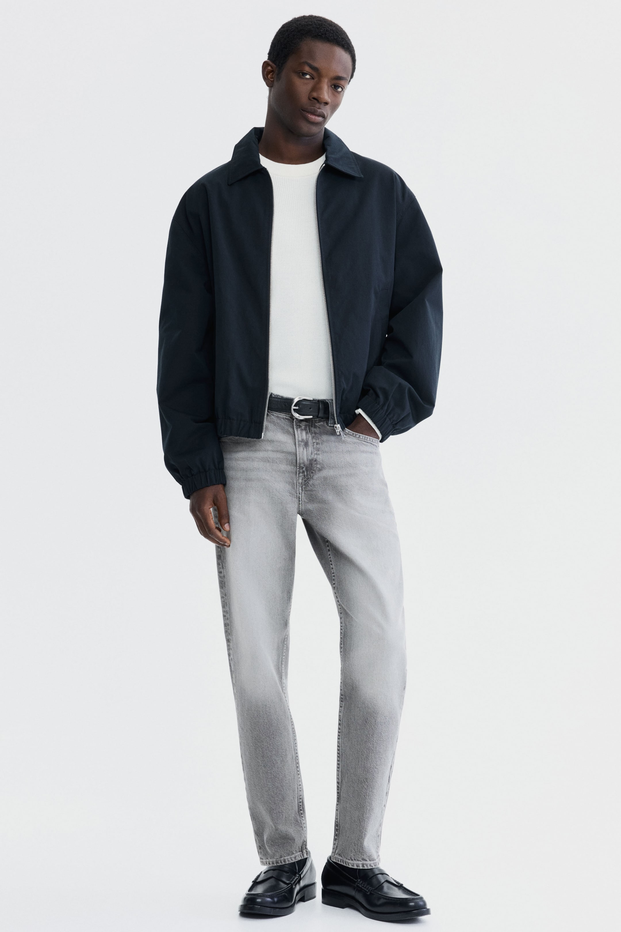 Visa större bild: Ljusgrå, urtvättade 5-ficksjeans med normal midja och avsmalnande ben bärs av modellen. Ett svart bälte håller jeansen på plats, och en av modellens händer vilar i en framficka.