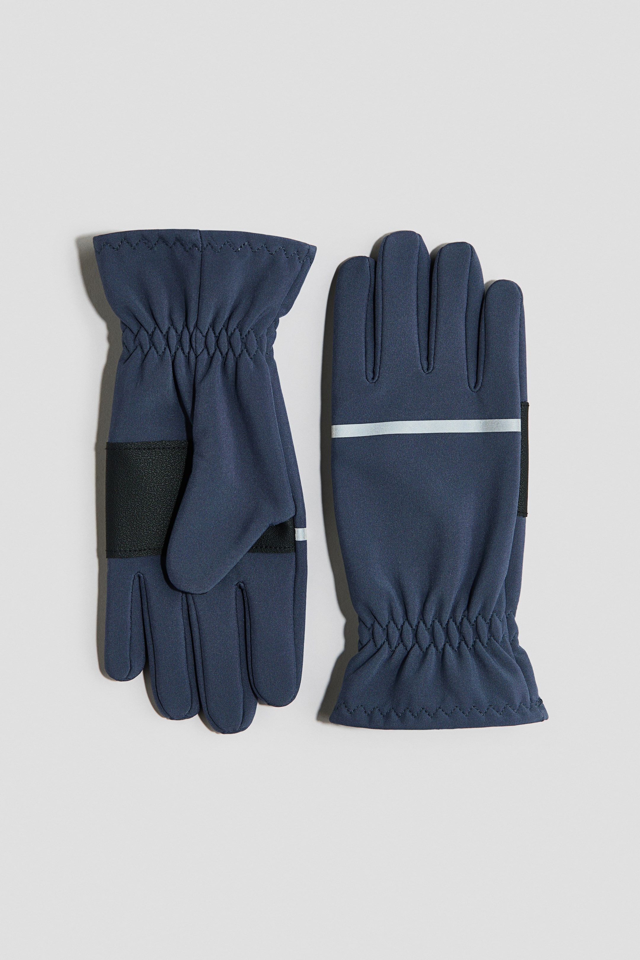 Ver imagen más grande: Un par de guantes azul marino está colocado en plano, mostrando el reverso. Ambos guantes presentan puños elásticos que crean frunces y dedos independientes. El guante izquierdo muestra una correa negra con una franja reflectante sobre el dorso de la mano, mientras que el guante derecho presenta una franja reflectante horizontal y una zona rectangular negra cerca de la muñeca.