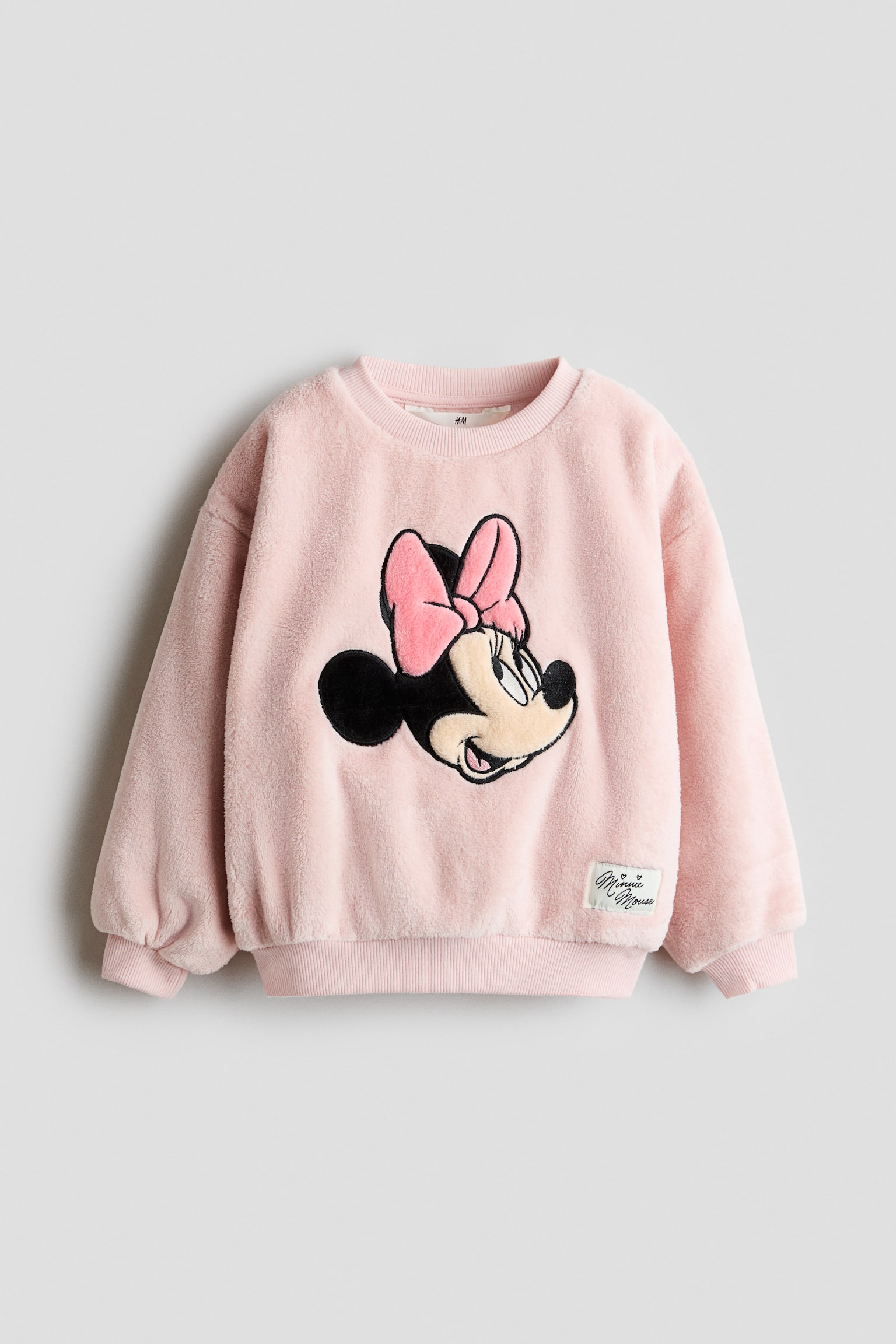 En ljusrosa, mjuk fleecetröja visas framifrån och har ett stort broderat Minnie Mouse-märke mitt på bröstet. Tröjan har en ribbad rund halsringning, nedhasade axlar med ribbade muddar, ribbad nederkant och en liten vit vävd etikett nära den nedre kanten.
