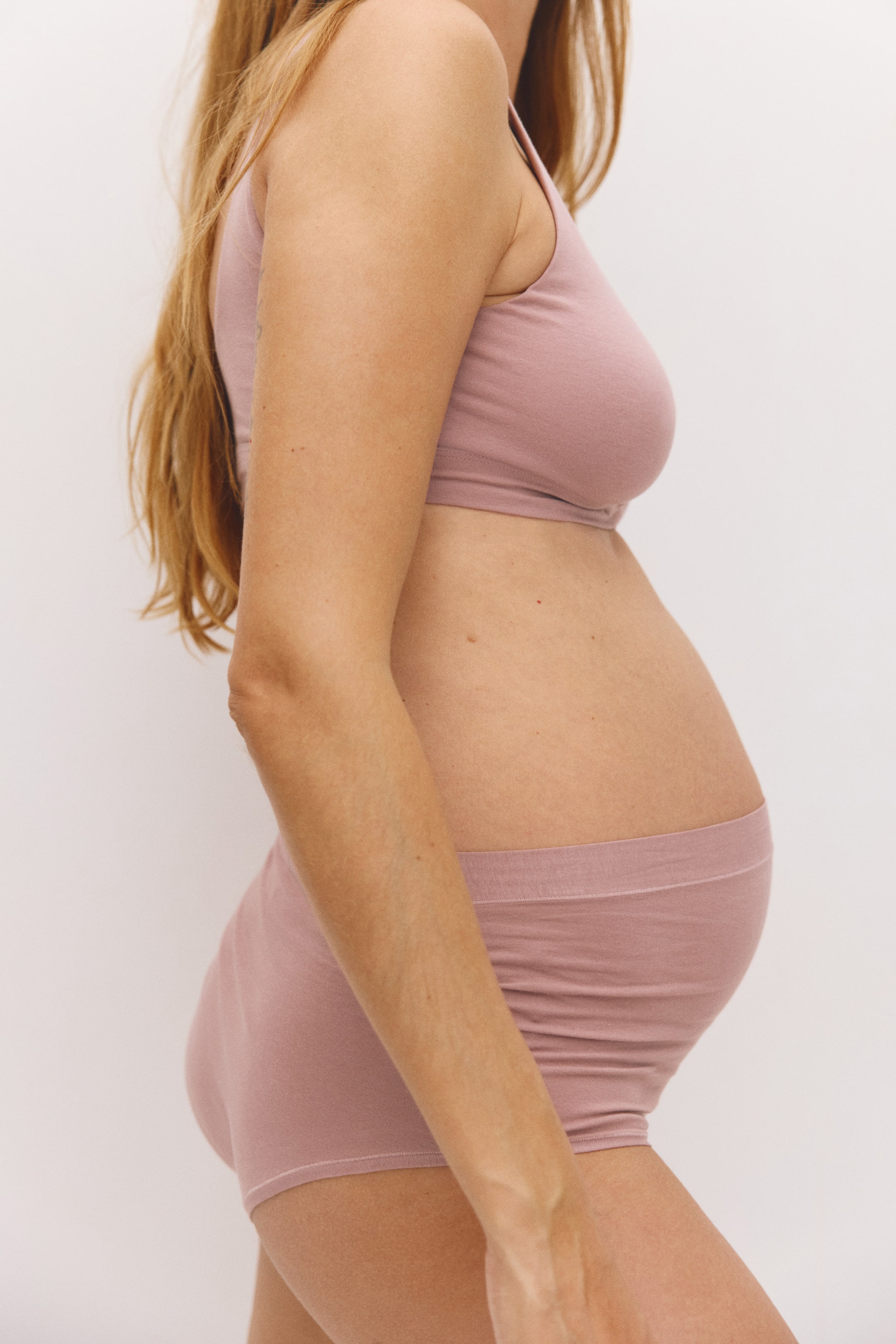 View larger image: The model is wearing high-waisted briefs in dusty pink cotton jersey, featuring side gathers that stretch over a pregnant tummy. The briefs are styled with a matching pink crop top.