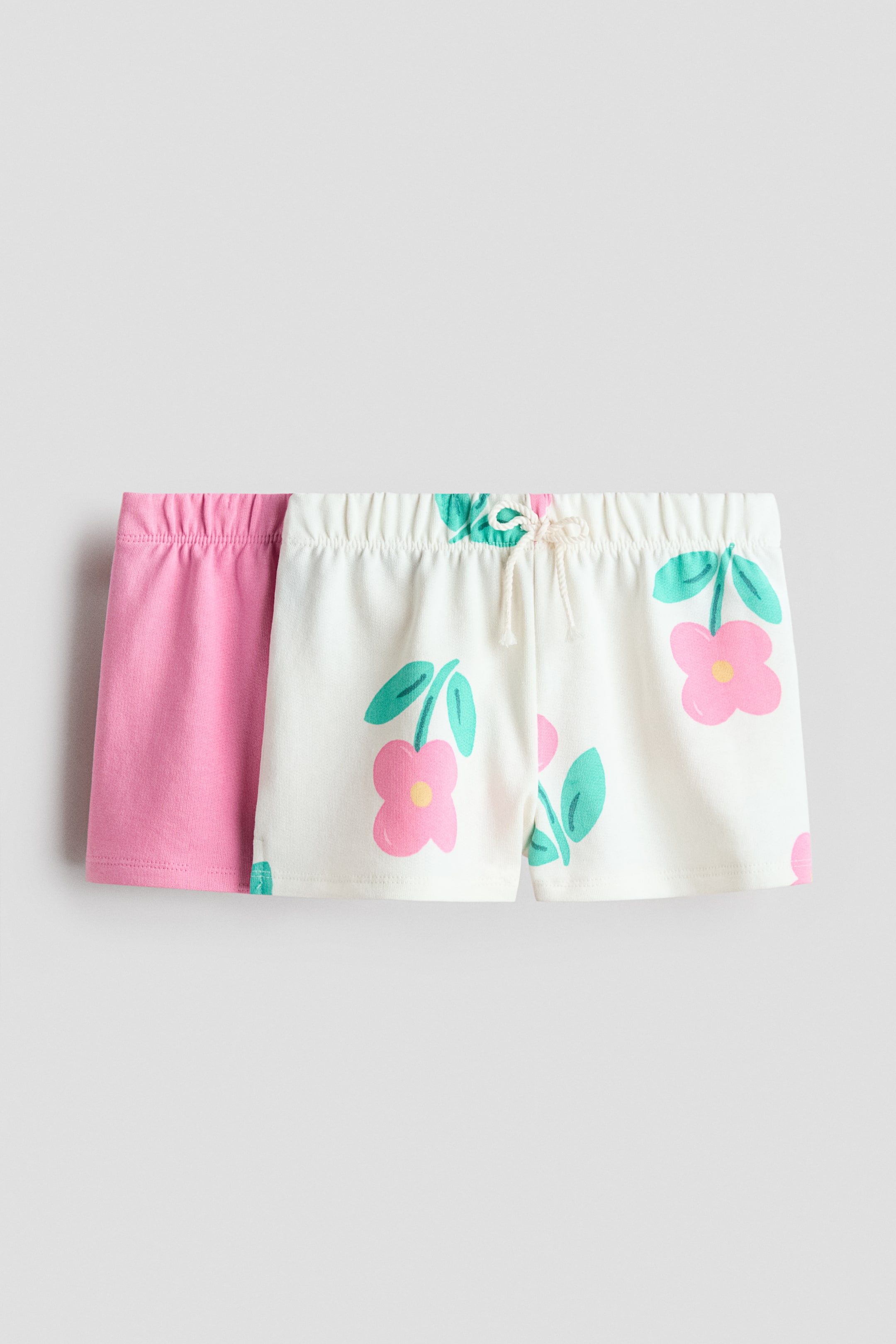 Grösseres Bild ansehen: Zwei Paar Shorts liegen flach, das eine leicht hinter dem anderen, und präsentieren ihre Vorderseiten. Das vordere Paar ist cremefarben und zeigt ein Blumenmuster mit rosa Blumen, gelben Zentren und türkisfarbenen Blättern. Es verfügt über einen elastischen Bund mit einer cremefarbenen Kordel, die vorne in der Mitte gebunden ist. Das zweite Paar, das dahinter sichtbar ist, ist einfarbig rosa und hat ebenfalls einen elastischen Bund.