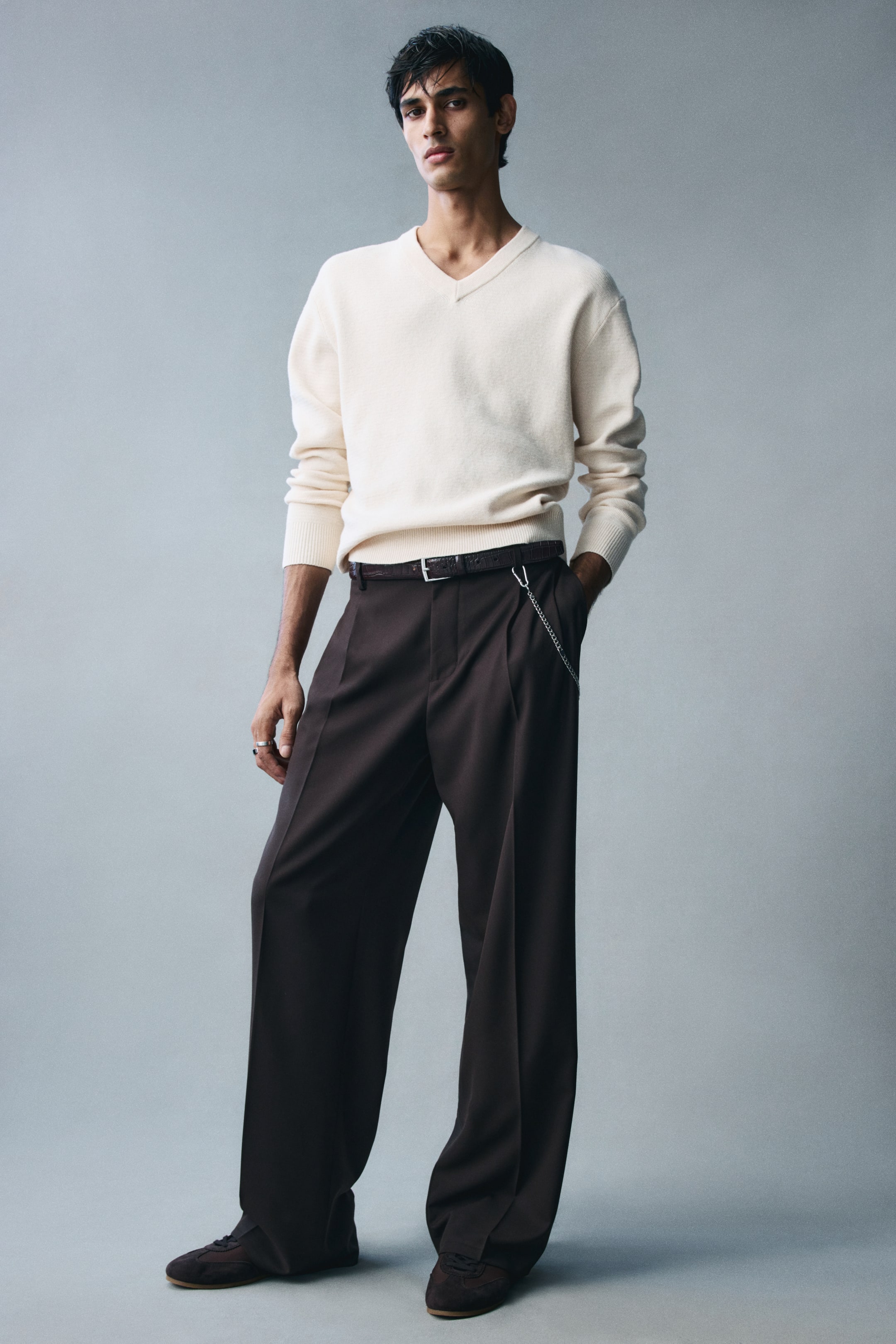 View larger image: A cream-coloured, cotton-blend knit sweater featuring a relaxed fit, a ribbed V-neck, and ribbed trim at the cuffs and hem is being worn by the model. The long-sleeved sweater is styled tucked into dark brown wide-leg trousers with a belt, with the sleeves slightly pushed up on the forearms.