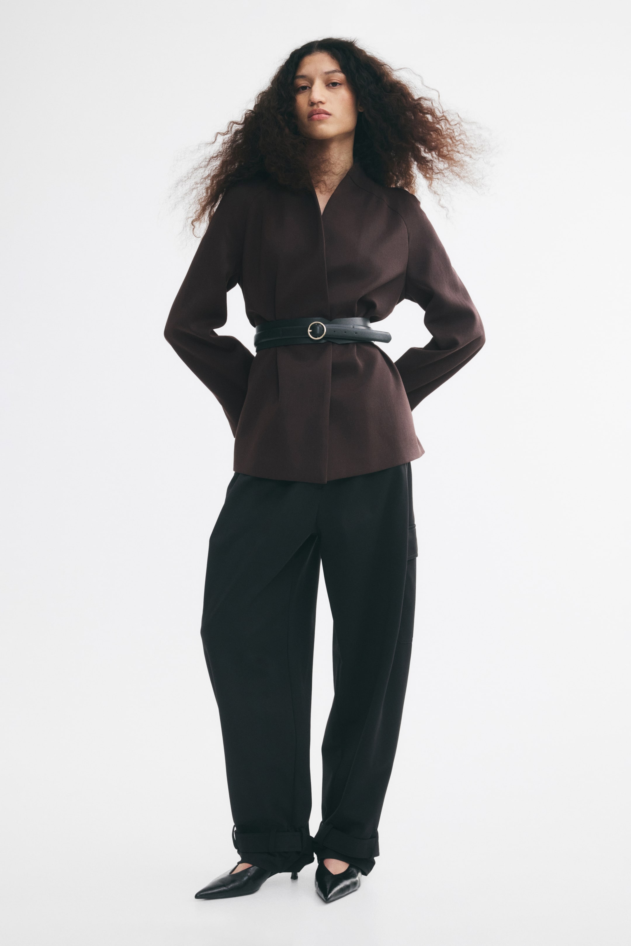 Größeres Bild ansehen: Ein dunkelbrauner, oversized geschnittener Blazer ohne Revers mit Wickelvorderteil und Sattelschultern wird vom Model getragen und in der Taille mit einem breiten, verstellbaren schwarzen Gürtel tailliert. Der Blazer ist mit locker geschnittenen schwarzen Hosen und schwarzen Schuhen mit spitzer Zehenpartie kombiniert.