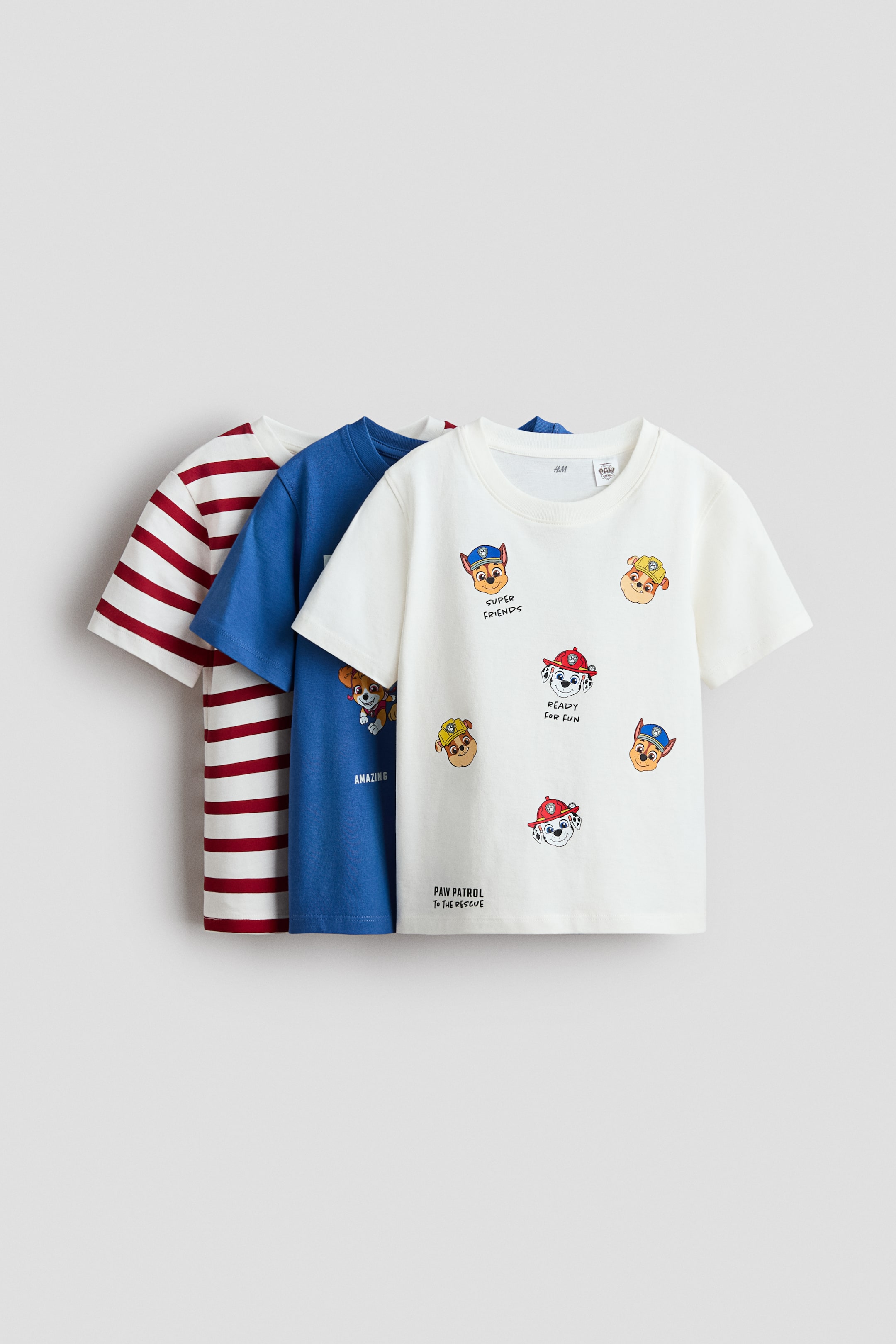 View larger image: Three classic short-sleeved T-shirts are on display, each with a rib-trimmed crew neckline. The front of a white cotton-jersey T-shirt is visible, featuring multiple colourful cartoon character prints and text, while a solid blue cotton-jersey T-shirt with a partial print and a red-and-white horizontally striped cotton-jersey T-shirt are partially visible behind it.