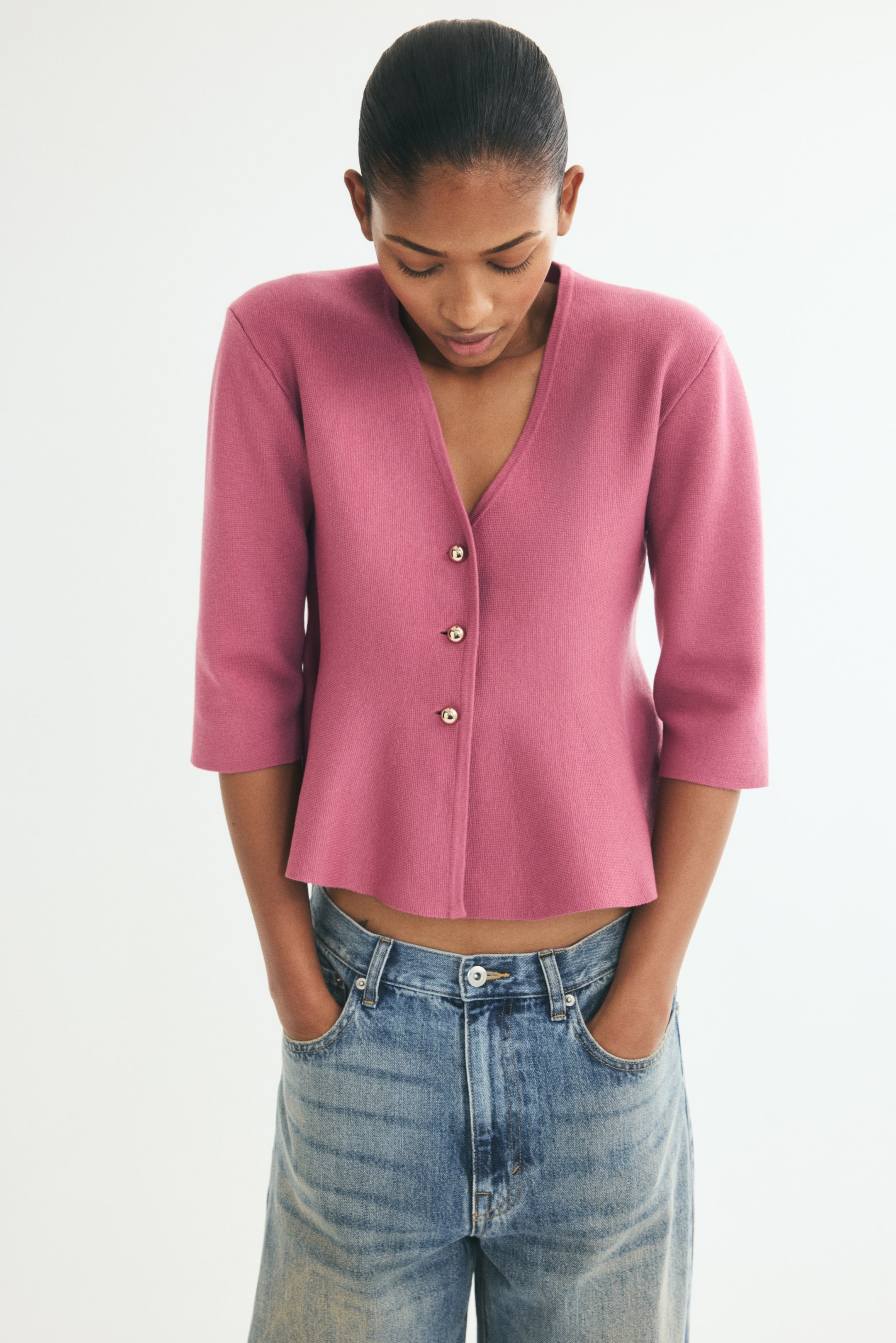 View larger image: A front-facing view shows a fitted pink cardigan in a high-density knit, featuring a V-shaped neckline and three gold-tone buttons down the front. The cardigan has gently dropped shoulders, 3/4-length sleeves, and a flared peplum hem, and is paired with blue jeans.