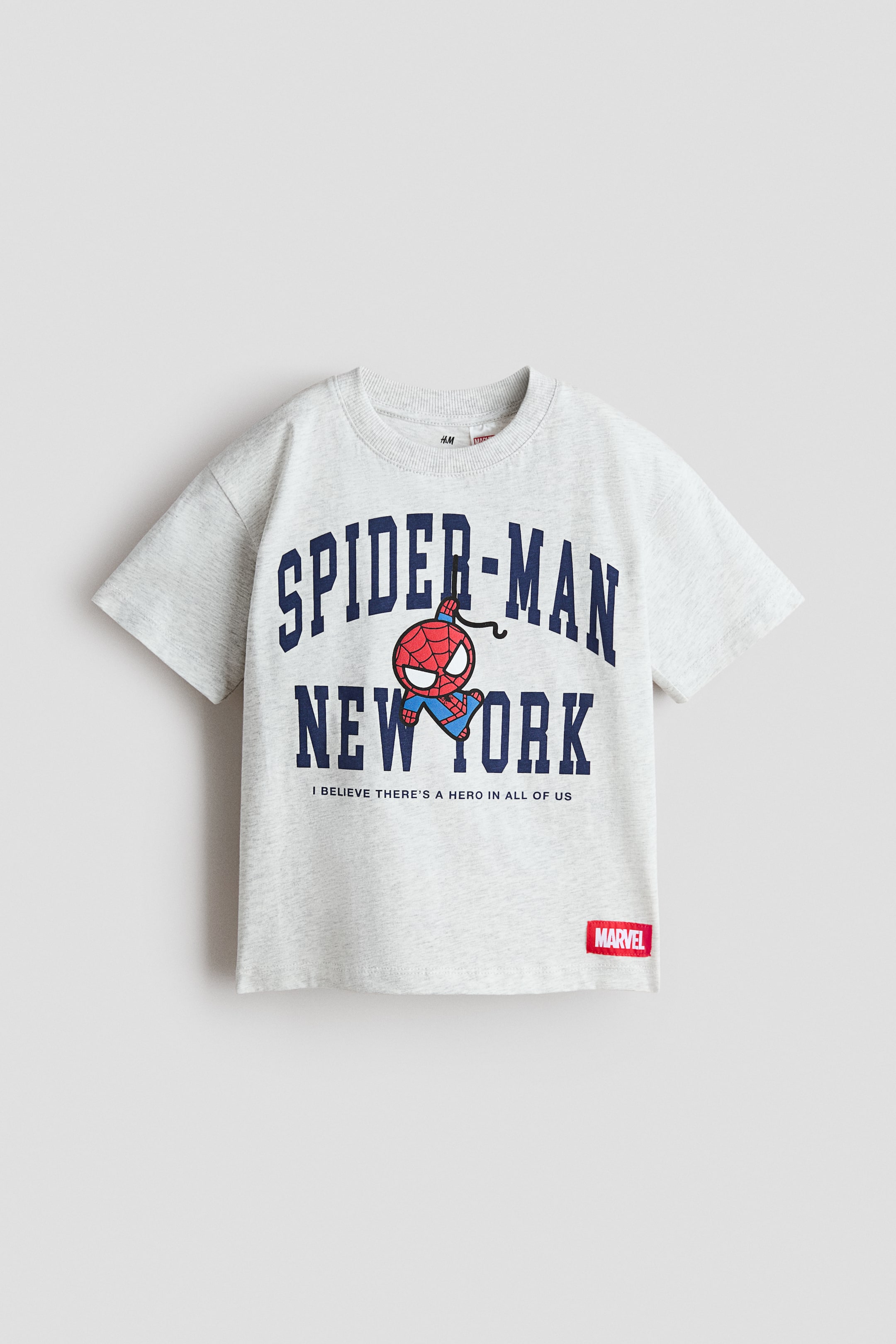 Agrandir l'image: T-shirt à coupe ample en jersey de coton doux gris clair à motifs imprimés, présenté de face. Il présente un col rond bordé de côtes et des épaules tombantes. Le devant affiche « SPIDER-MAN » et « NEW YORK » en lettrage universitaire bleu foncé, un petit personnage cartoon de Spider-Man ainsi que le texte « I BELIEVE THERE'S A HERO IN ALL OF US », avec une étiquette rouge « MARVEL » visible sur l'ourlet inférieur droit.